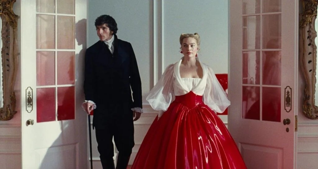 A person in a suit stands beside another in a flowing red gown and sheer shawl, entering through ornate doors in an elegant setting