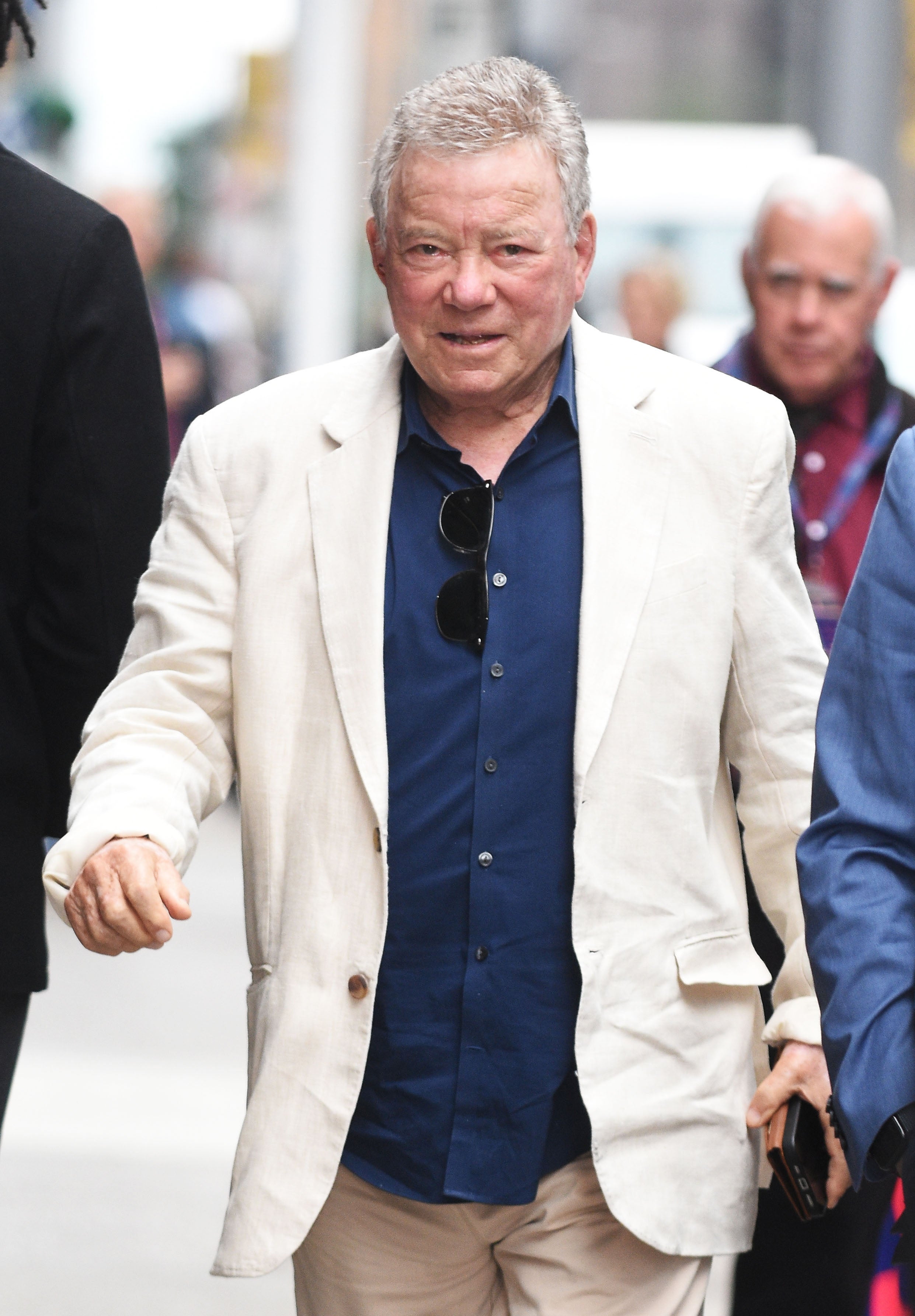 Person in beige suit with a dark shirt walks outdoors, holding sunglasses. They're engaged in conversation with others nearby