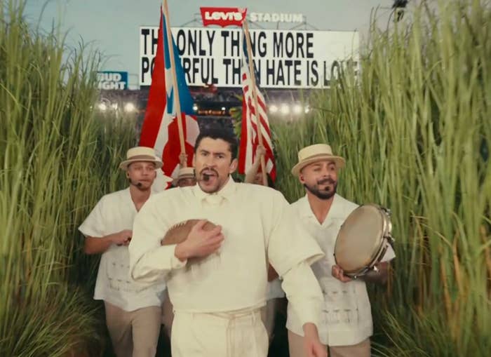 Grupo de hombres en ropa tradicional, uno toca güiro, delante de un cartel que dice: "The only thing more powerful than hate is love."