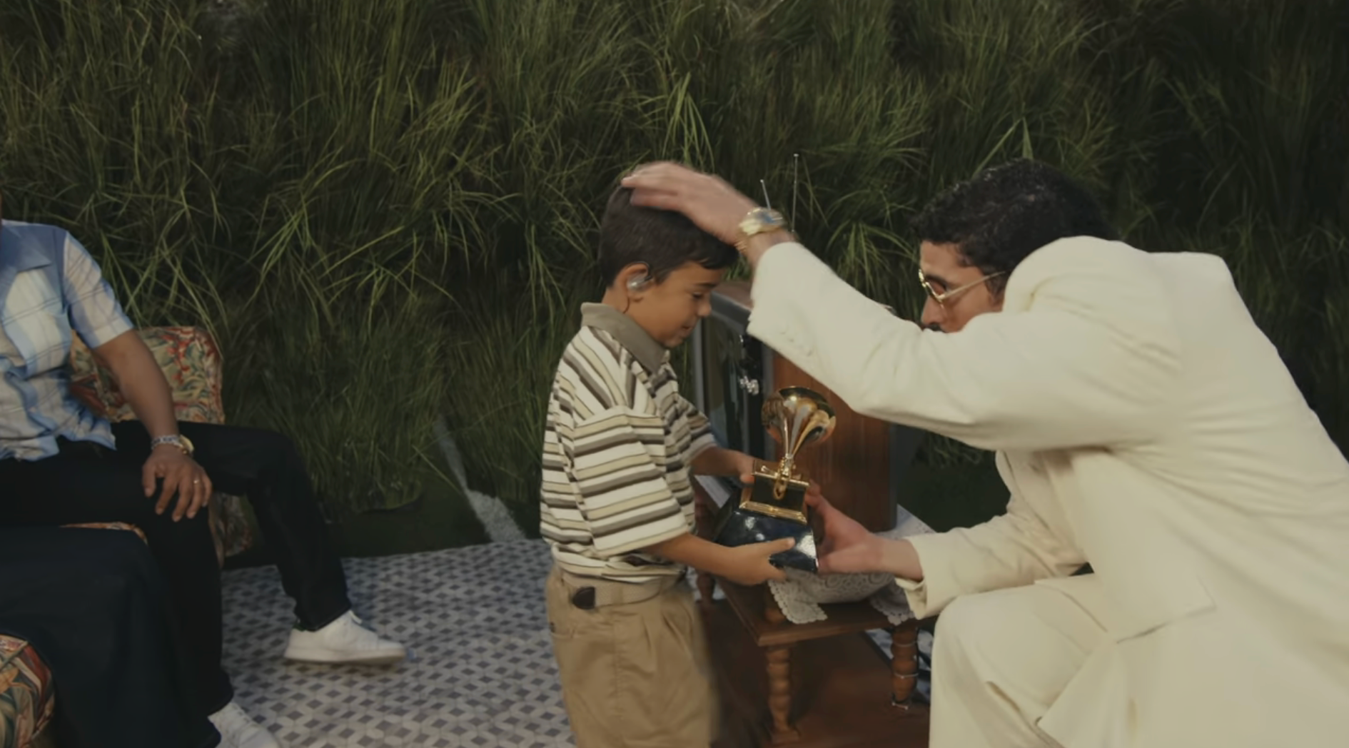 An big  successful  a suit   presents a young lad  with a trophy, some  smiling. Grass background, seated radical   partially visible