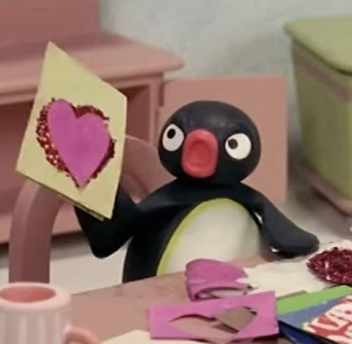 Pingüino de plastilina sostiene una tarjeta artesanal con un corazón recortado. 