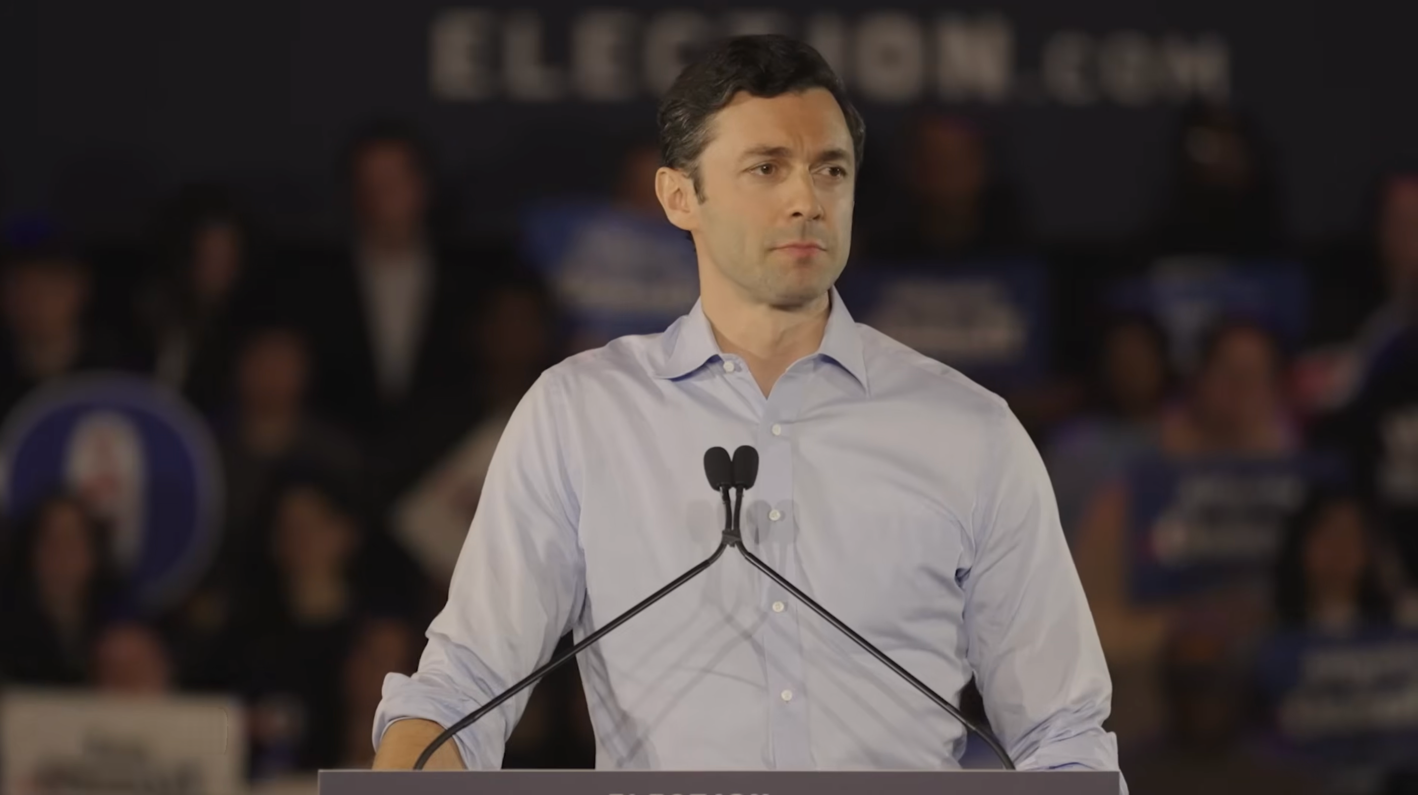 A idiosyncratic   successful  a button-up garment  speaks astatine  a podium during an predetermination  event, with a blurred assemblage  and signs successful  the background