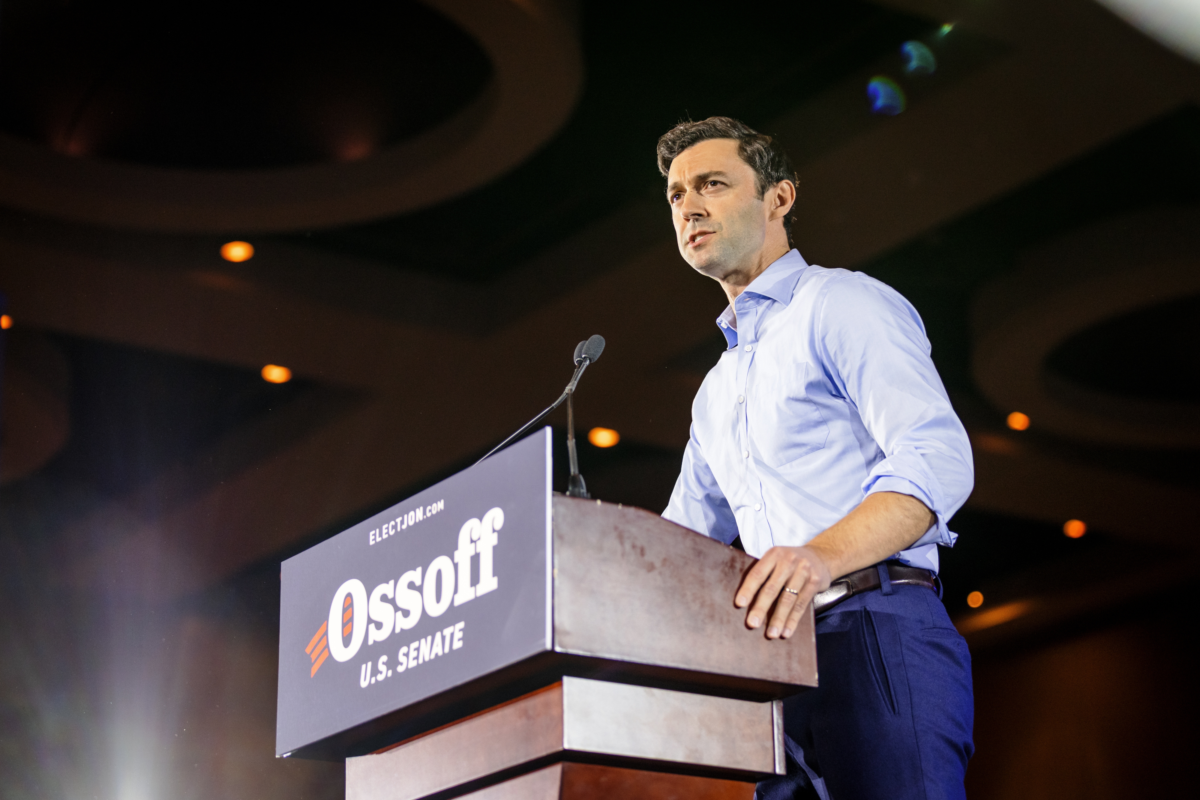 Person speaking astatine  a podium with "Elect Jon Ossoff U.S. Senate" sign, wearing a collared garment  and pants, addressing an assemblage  indoors
