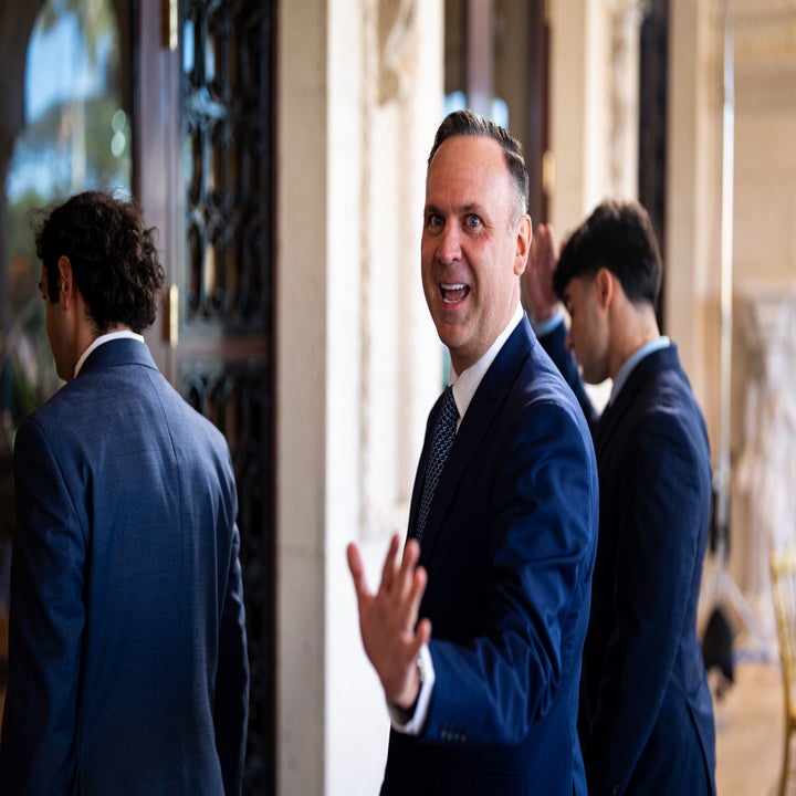 Dan Scavino waves with a smile, standing near others in suits outside a building