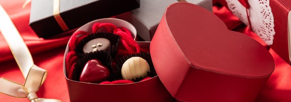 赤いハート形のボックスにチョコレートが詰められたギフト。背景に他のギフトボックスとリボンが並ぶ。