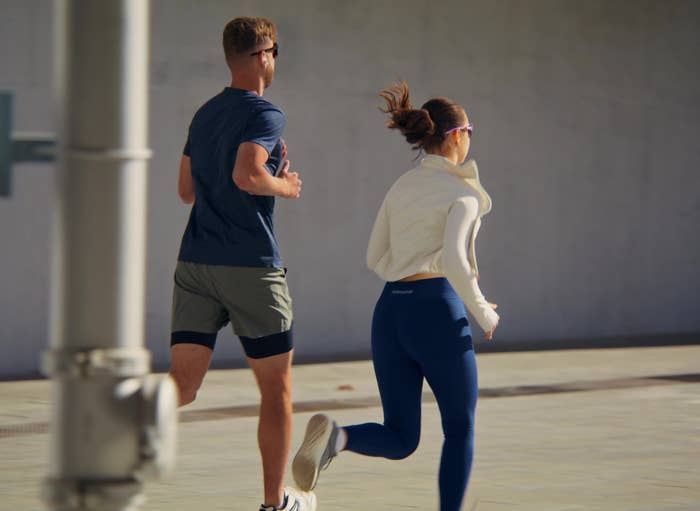 Two people jogging outdoors on a sunlit pavement, wearing athletic clothing; one in shorts and the other in leggings