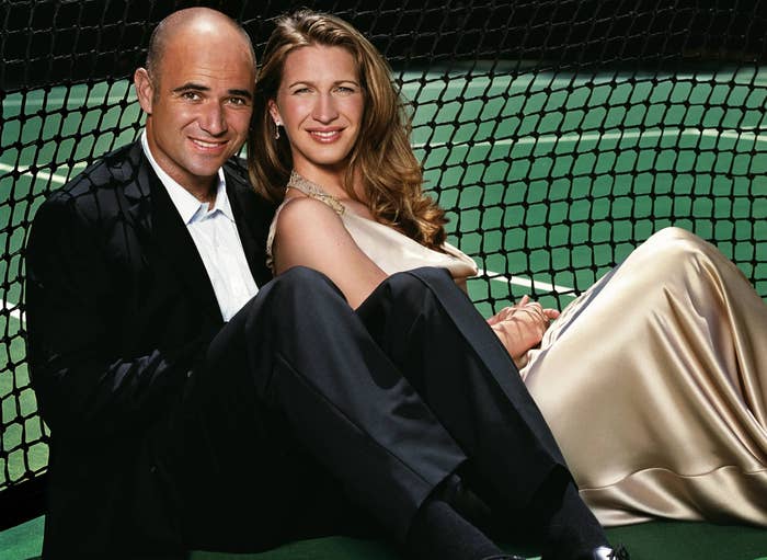 Two individuals sit on a tennis court, smiling at the camera. The woman wears an elegant gown, while the man is dressed in a suit