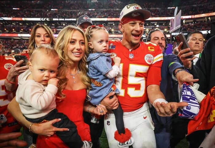 Family celebrates on the field, holding children, after a football game. One adult wears a football jersey, another is in a red dress