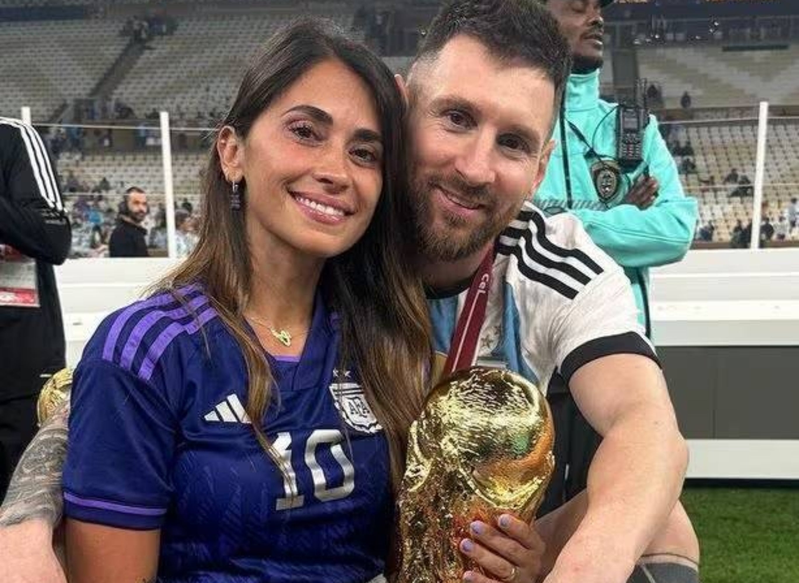 A man and woman sit together in a stadium, holding a trophy. She wears a sports jersey; he wears a uniform with a medal