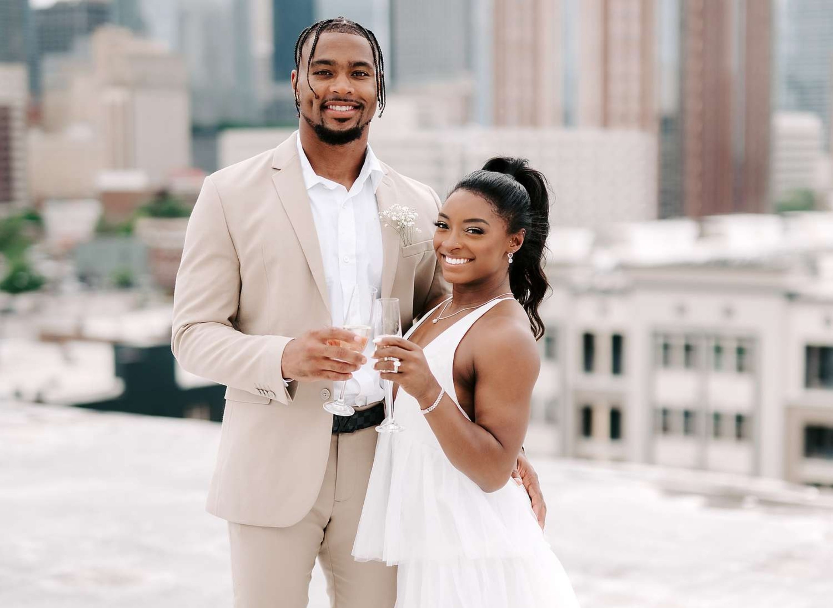 A couple on a rooftop, dressed elegantly. The man wears a suit, the woman a tiered dress. They hold champagne, smiling against a city backdrop