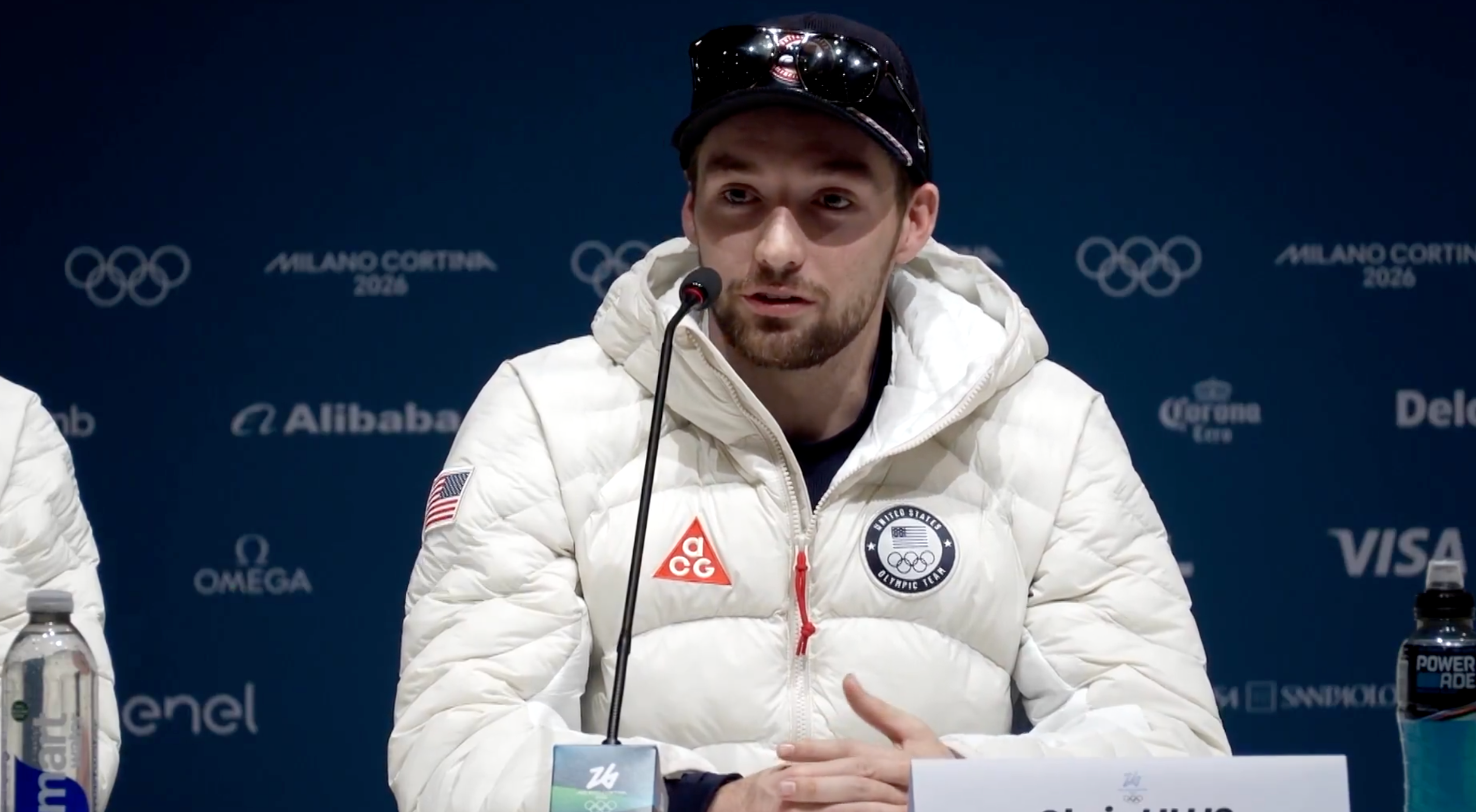 Athlete astatine  property   league  successful  a branded overgarment   with logos speaks into a microphone, wearing a headdress  and sunglasses, against a backdrop of sponsor logos