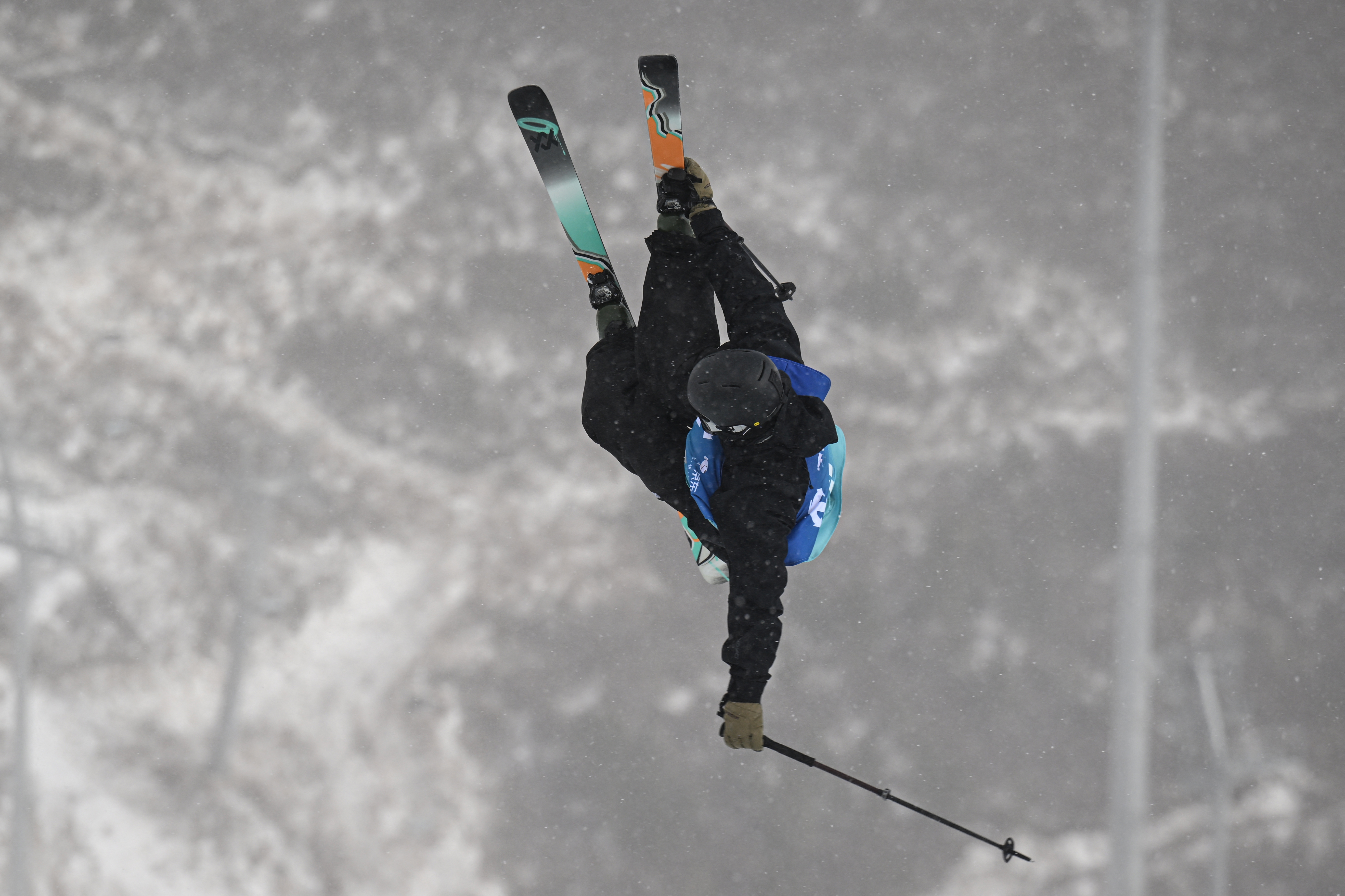 Skier performs a high-flying aerial instrumentality   with skis crossed, wearing a helmet and snowfall  gear, against a snowy mountainous backdrop