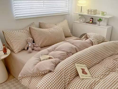 Habitación acogedora con cama desordenada de cuadros, peluche y libro en la colcha. Mesita con lámpara y tazas en estante al fondo