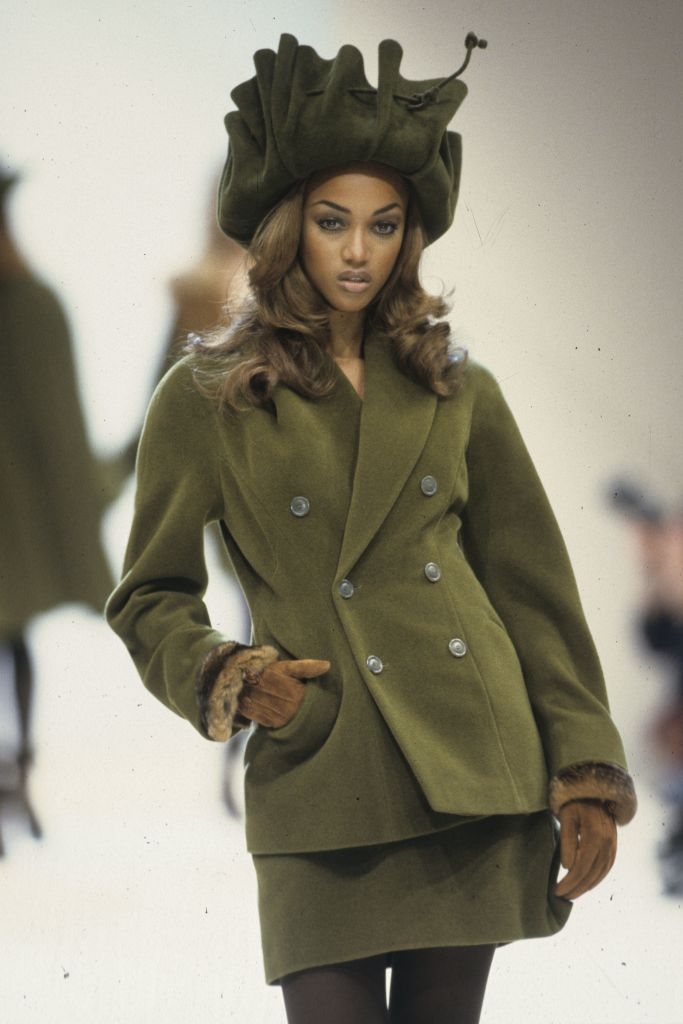 Model successful  stylish outfit with a structured coat, skirt, gloves, and a unsocial   hat, walking down   the runway