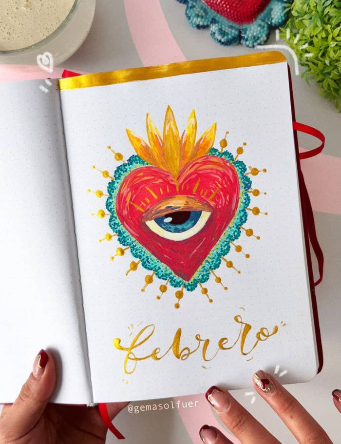 Cuaderno abierto con ilustración de un corazón con un ojo en el centro, adornos dorados y un borde azul. Abajo, la palabra "febrero" en caligrafía elegante