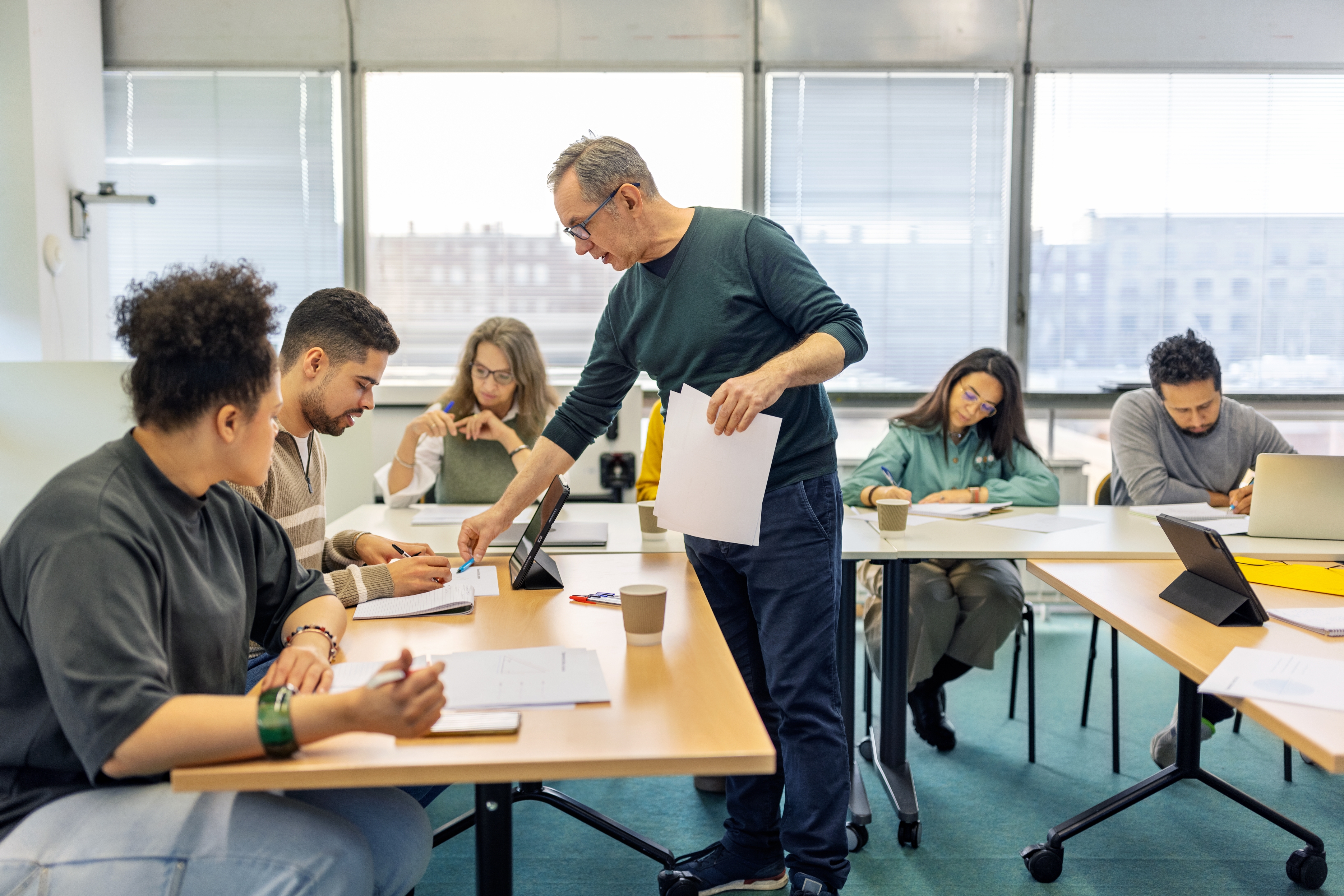 A group of people in a classroom setting, working at tables with laptops and papers, while an instructor assists them