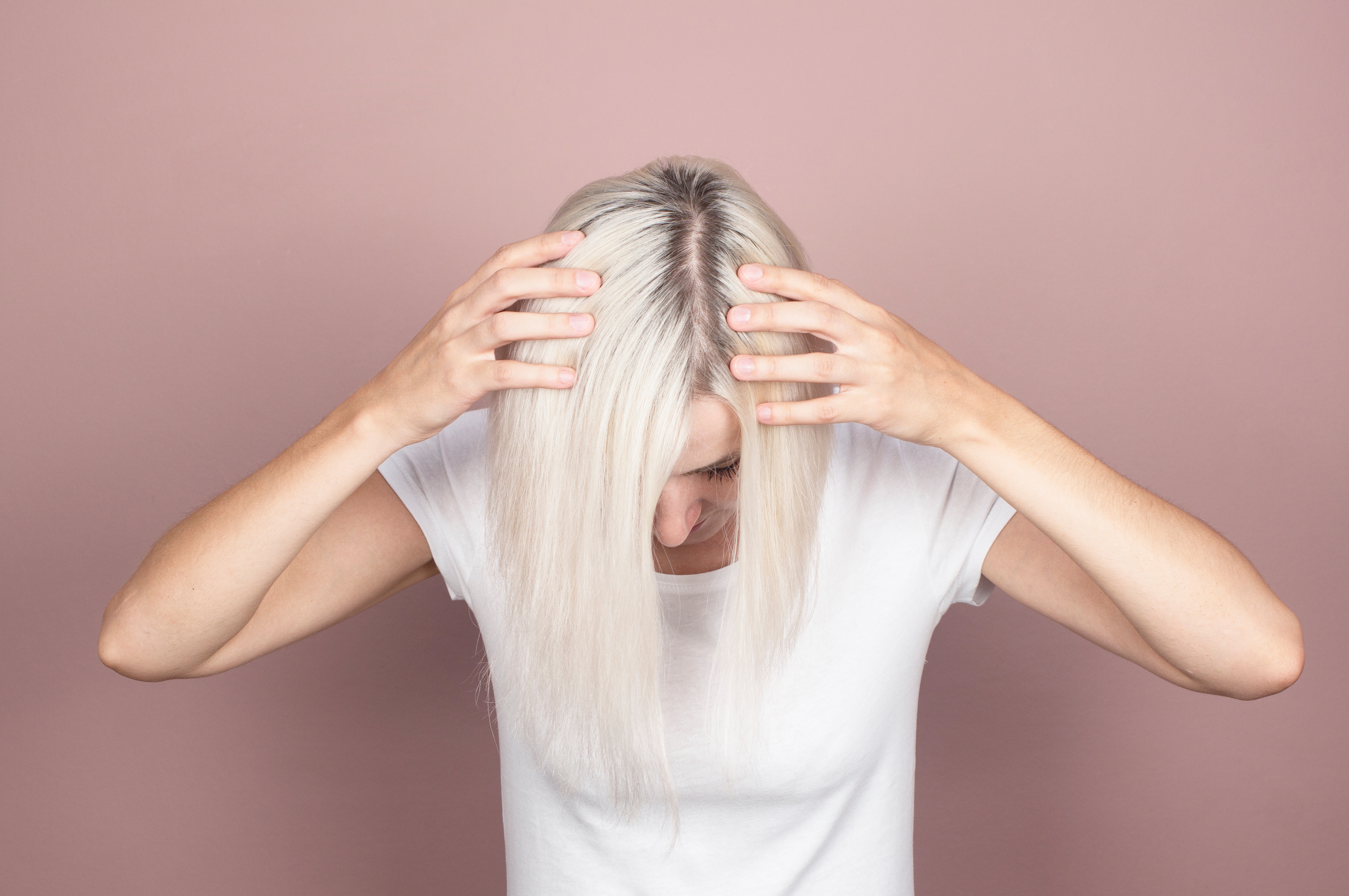Person with light hair parting it to reveal roots against a plain background, wearing a simple white T-shirt, looking down