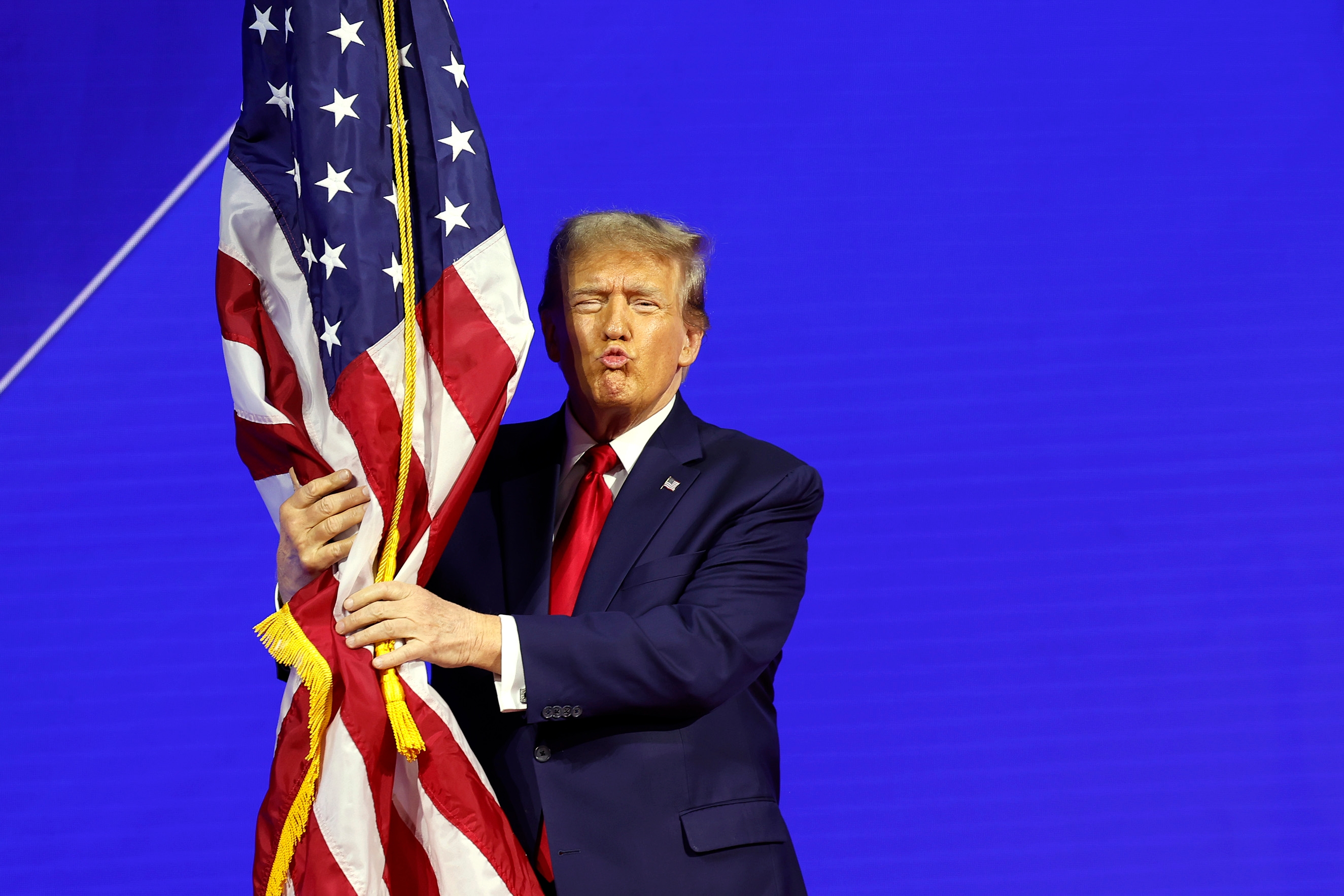 Person in a suit embraces and puckers lips at a large American flag on stage