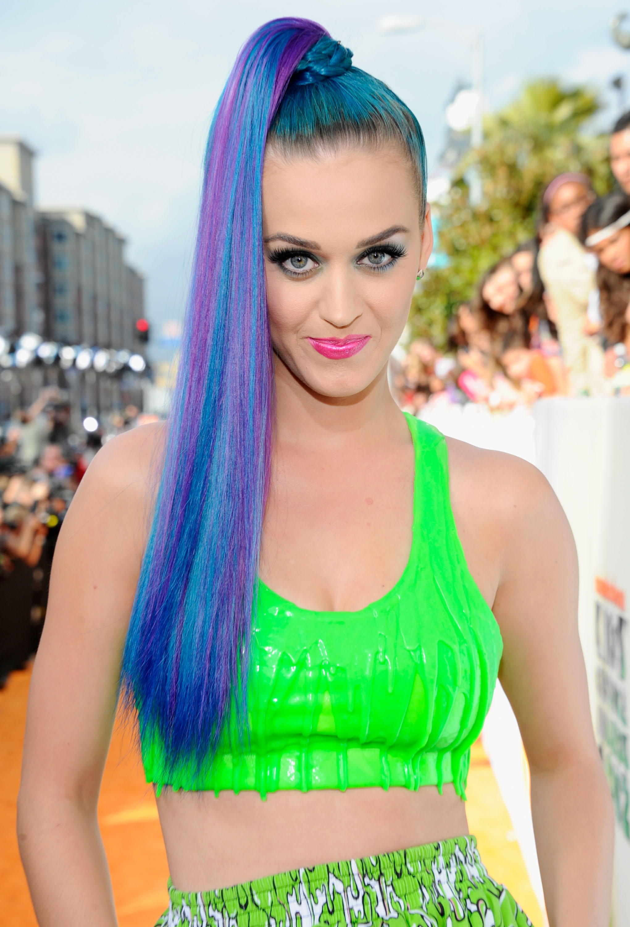 Person in green cropped top with structured style and multicolored high ponytail on an event-like setting. Peers and photographers visible in background