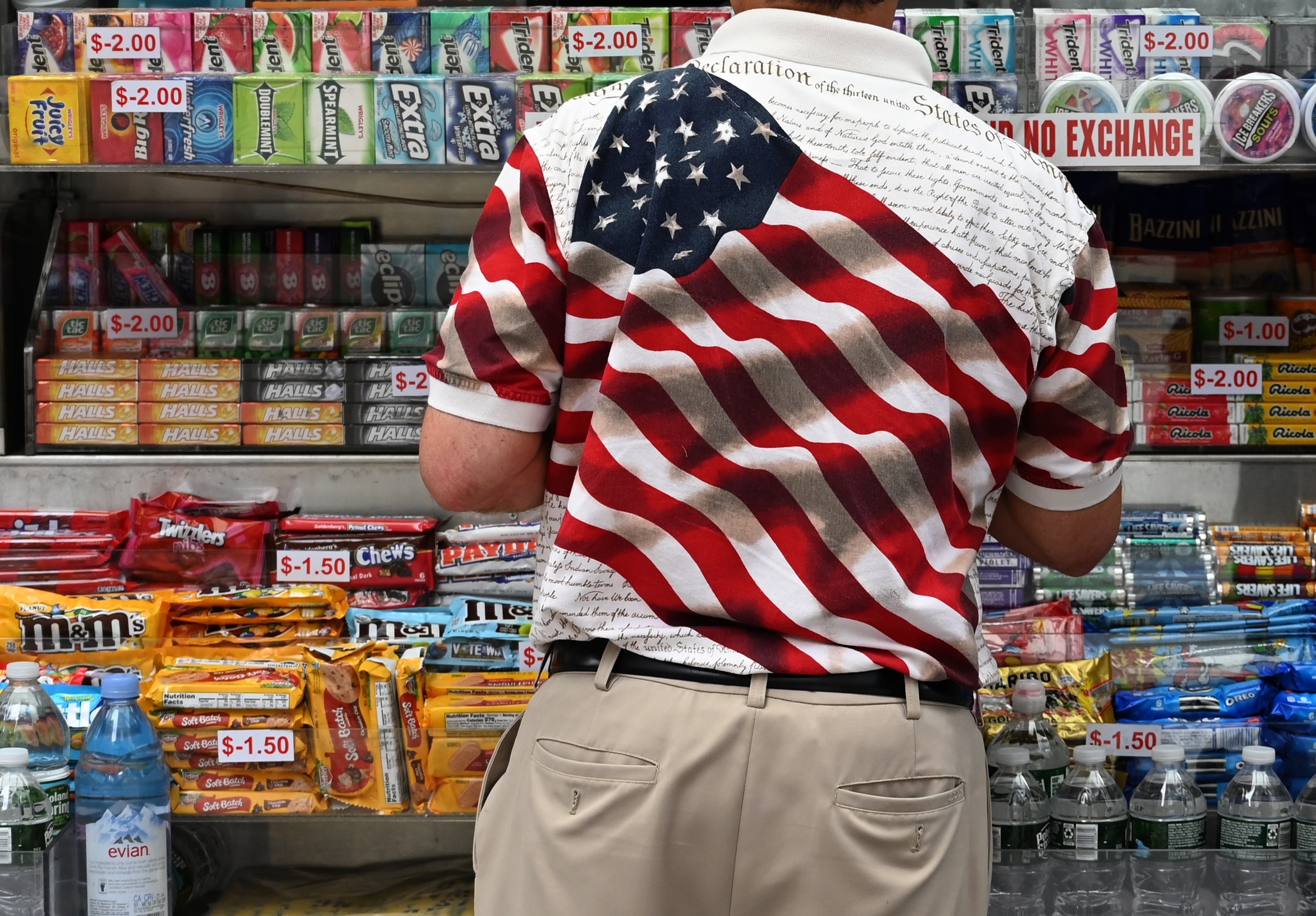 Person wearing a patriotic shirt with the U.S. flag design, standing in front of a candy and soda stand displaying various items and prices