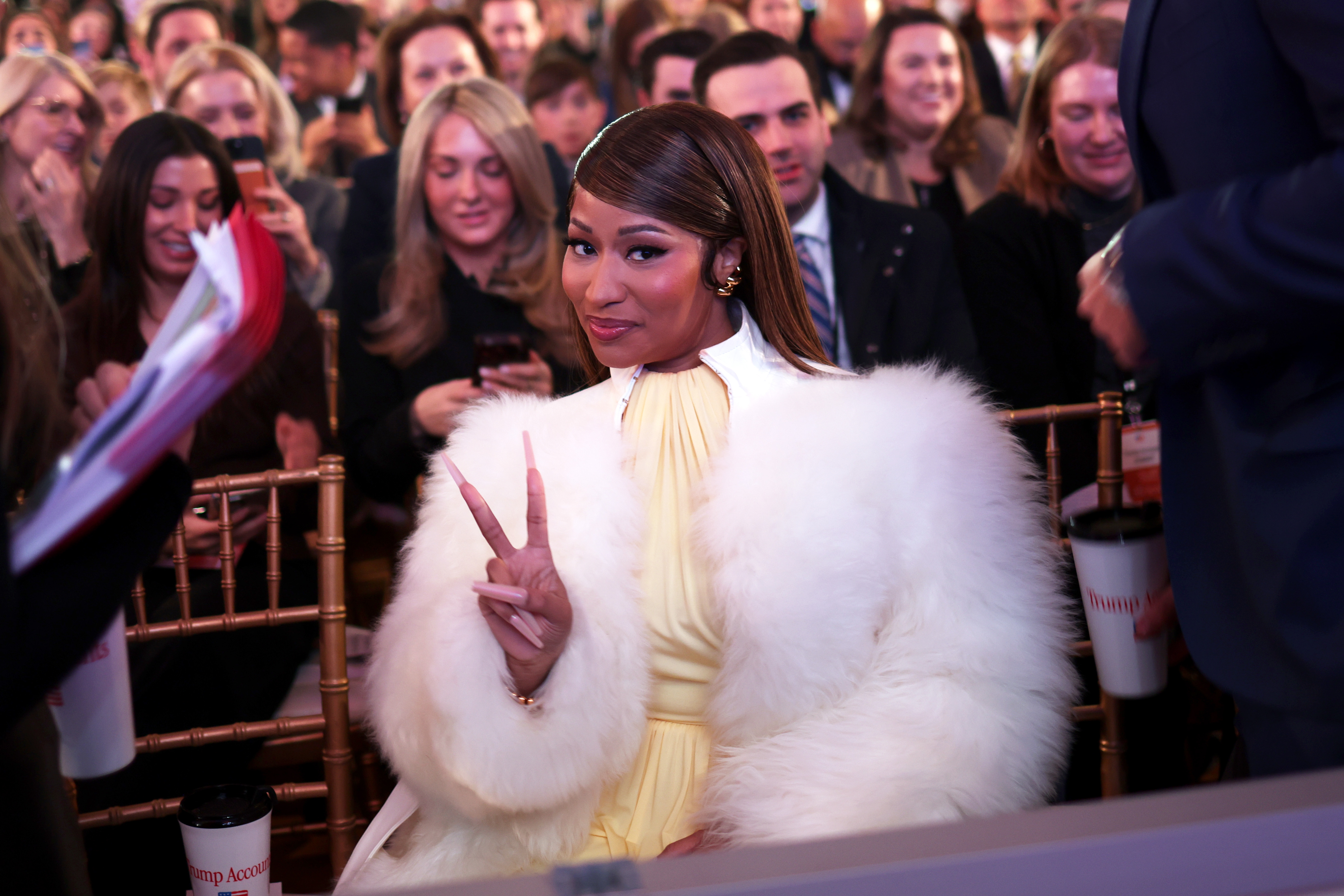 Person wearing elegant white fur coat and yellow dress, seated among audience, smiling and flashing peace sign with fingers