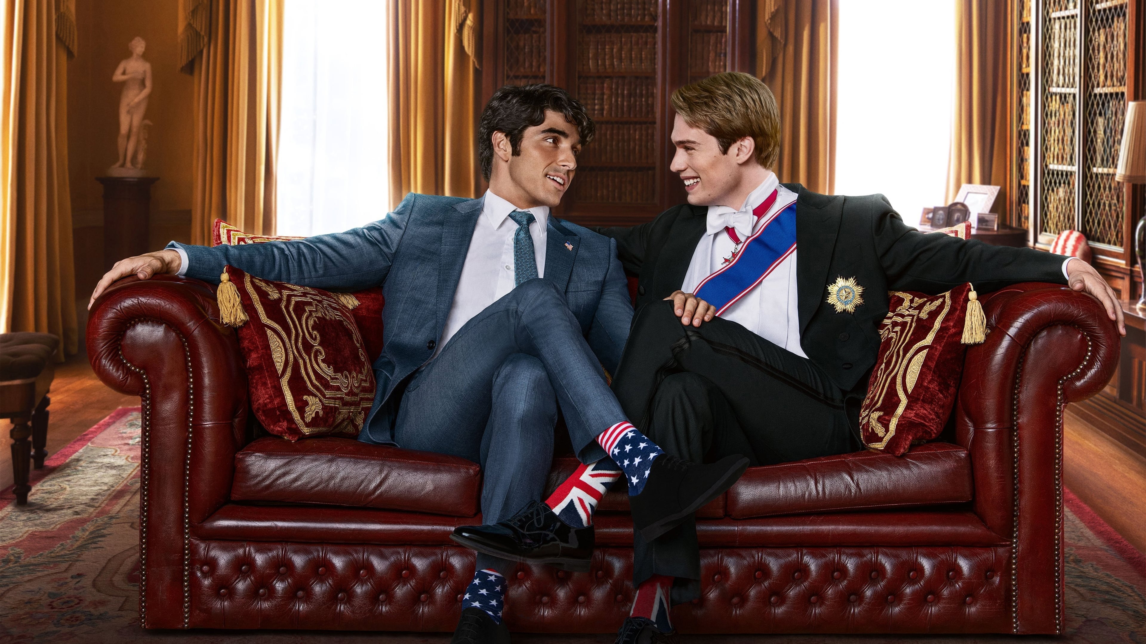 Dos personas en un sofá de cuero, una en traje azul con calcetines de bandera británica y otra con frac y banda ceremonial