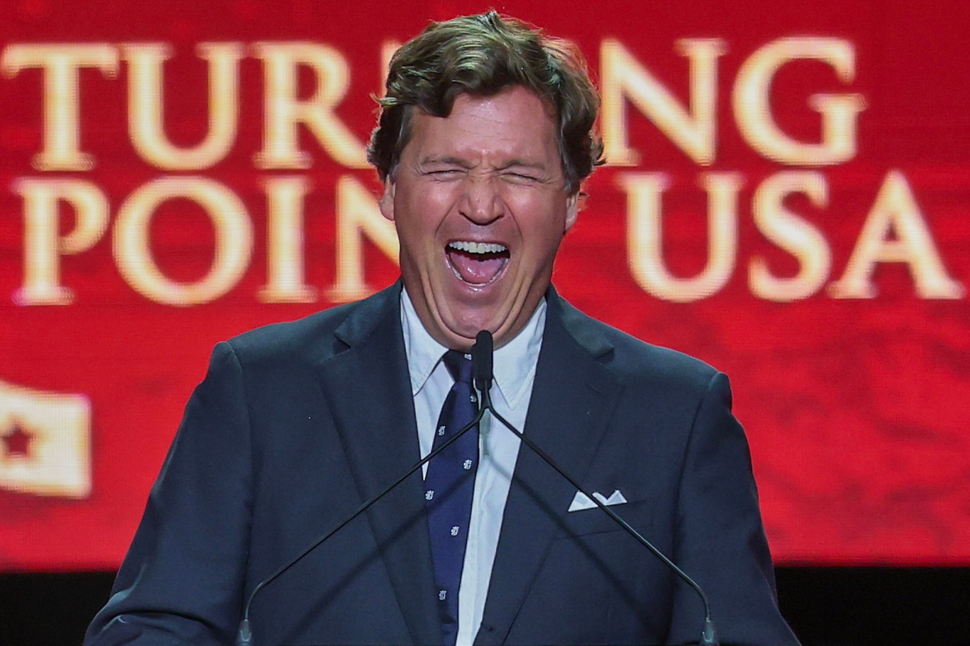 Person in a suit laughing at a podium in front of a Turning Point USA backdrop