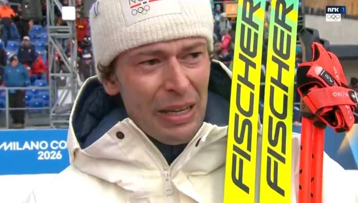 Person successful  wintertime  sports gear, with skis labeled "Fischer," appears affectional  astatine  an event. Background shows "Milano Cortina 2026" signage