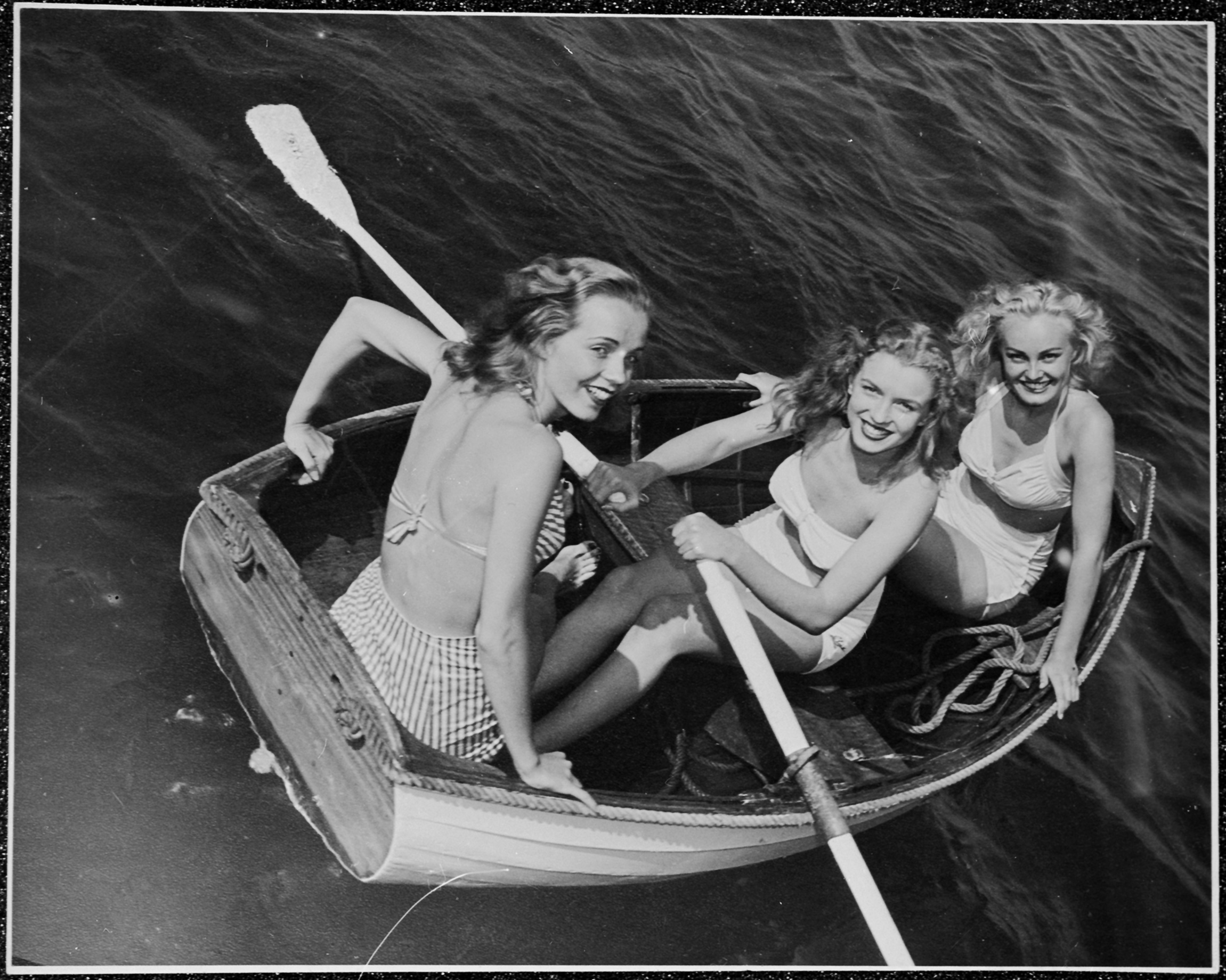 Three women successful  a tiny  rowboat, smiling and posing for the camera. They deterioration  swimsuits from a vintage era