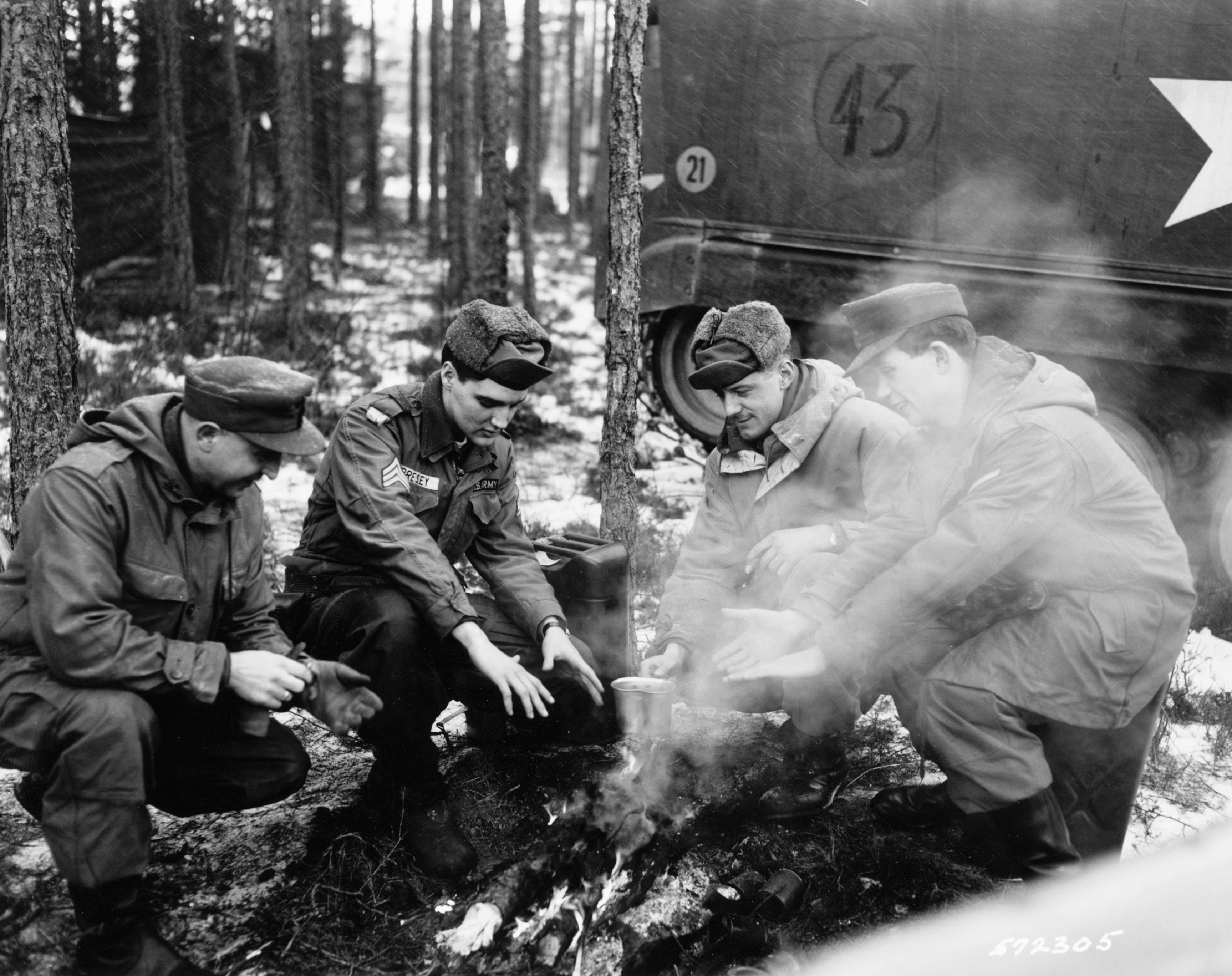 Four soldiers successful  wintertime  cogwheel  stitchery  astir   a tiny  campfire successful  a forested area, warming their hands. A subject   conveyance  is partially visible