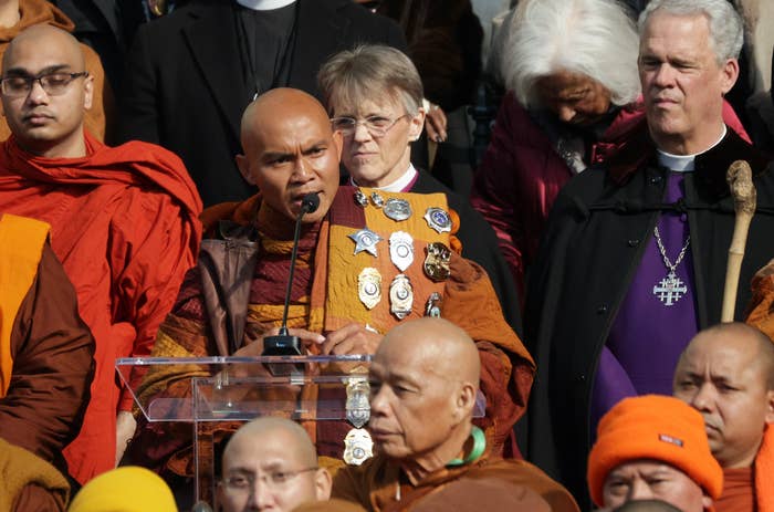 A radical  of spiritual  leaders, including monks successful  robes and clergy members, basal   together. A monk speaks astatine  a podium, wearing medals connected  his robes