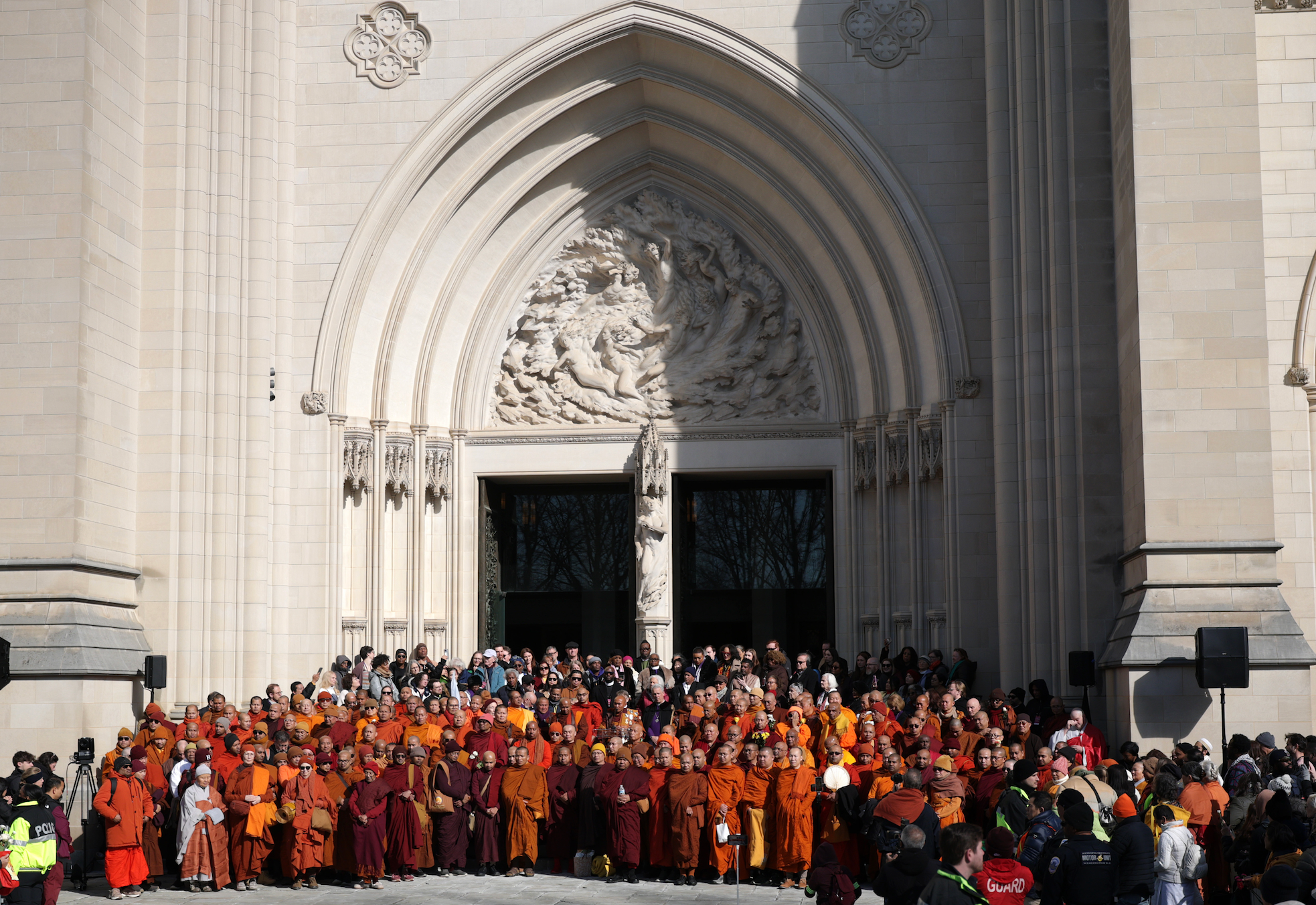 Para biksu berjubah tradisional berkumpul di depan katedral besar untuk sebuah acara, dengan ukiran batu yang rumit di atas pintu masuk