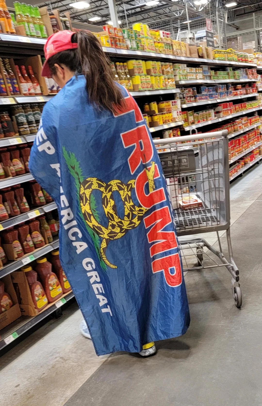 Person wearing a "Trump Keep America Great" cape successful  a market  store   adjacent  to a buying  cart
