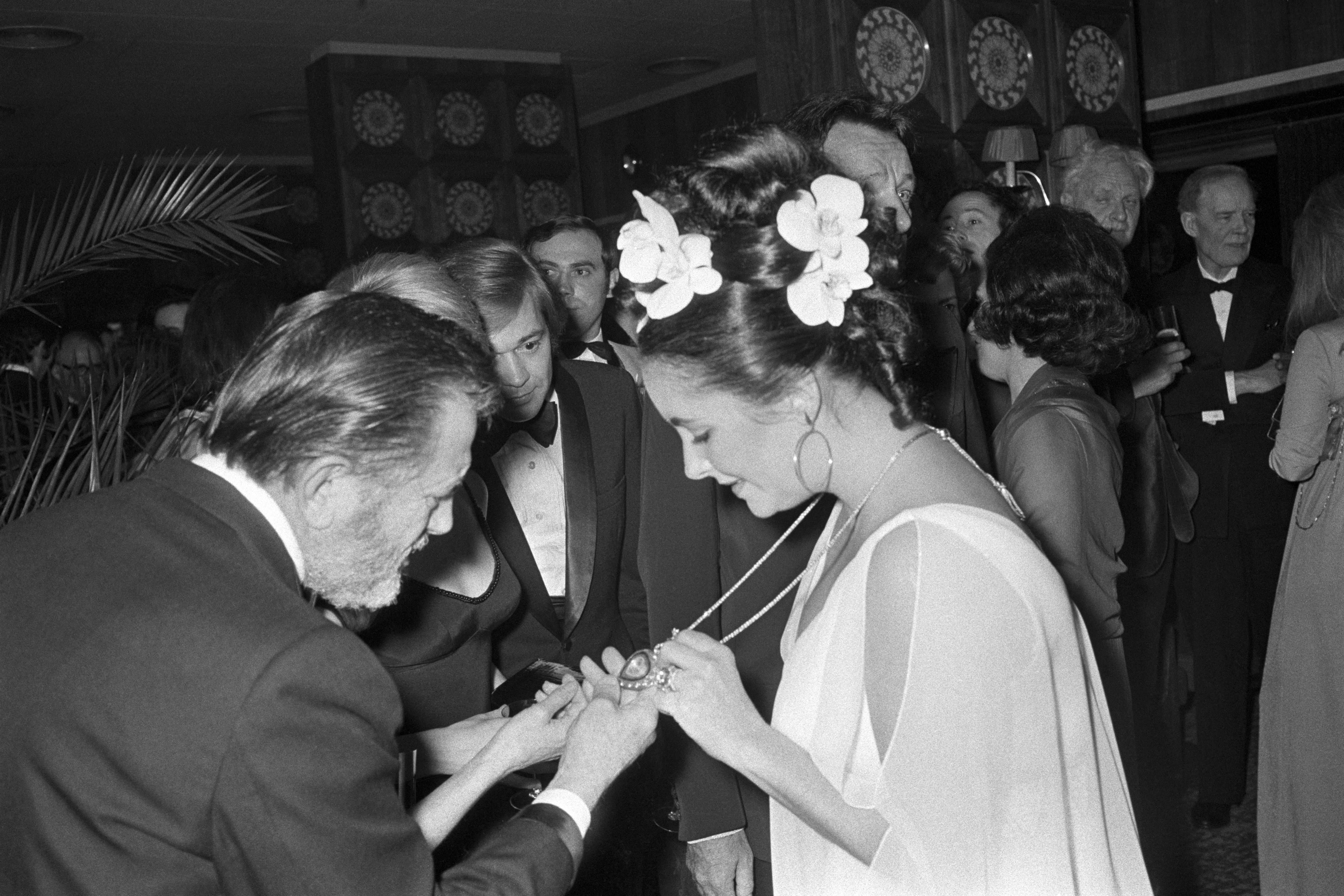 Woman successful  a stylish gown with floral hairsbreadth  accessories shows her necklace to a antheral   astatine  an elegant event. Guests successful  ceremonial  attire successful  the background