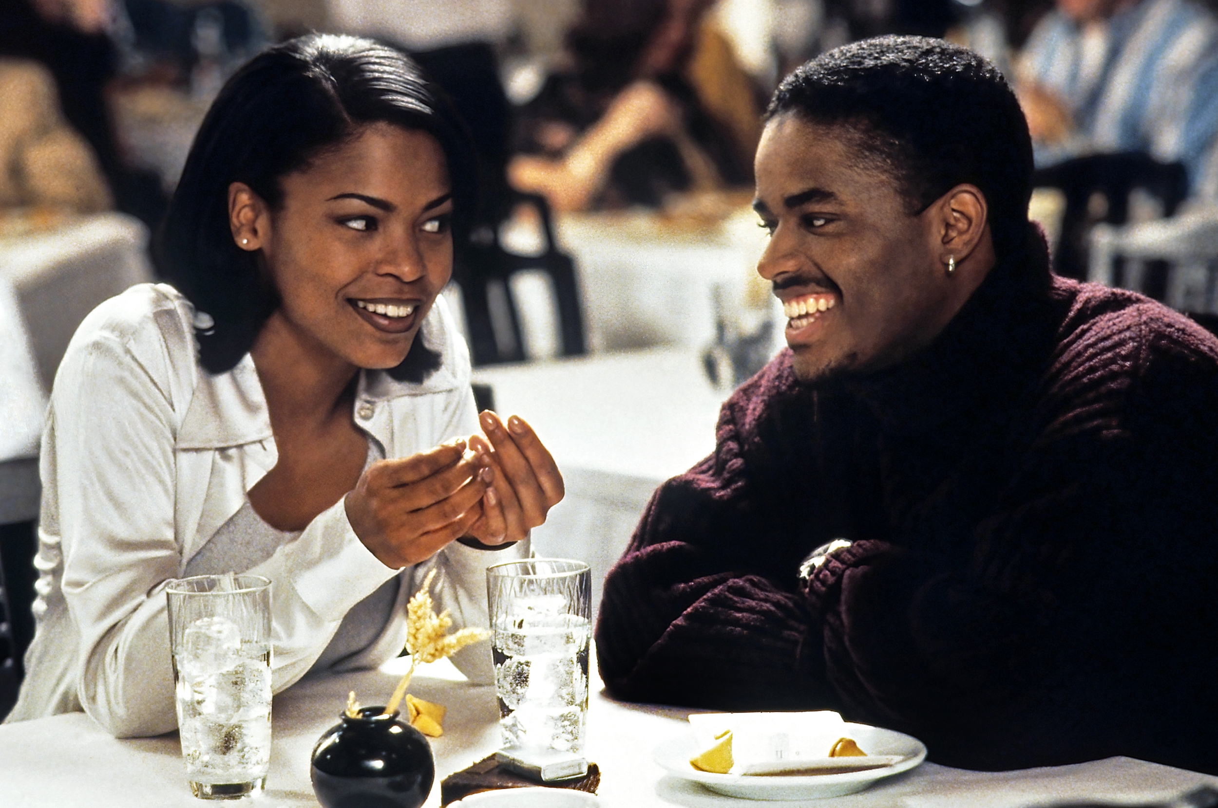 Two people sit at a table, smiling warmly at each other, with drinks in front of them in a relaxed setting