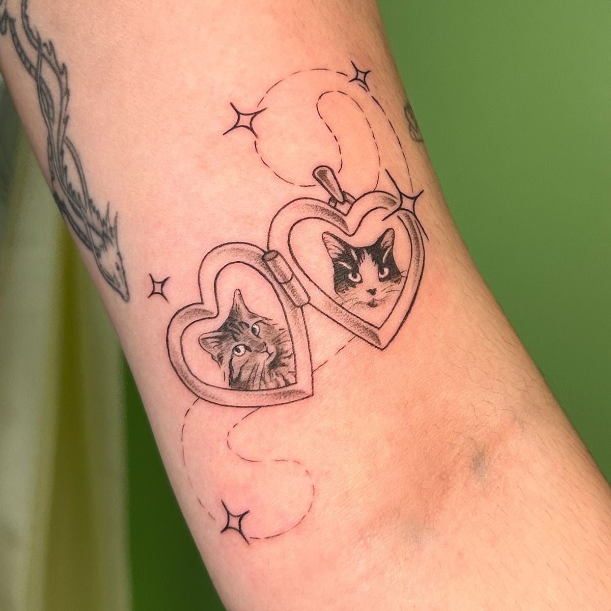 Tatuaje en el brazo muestra dos gatos en un relicario con forma de corazón, rodeado de líneas y estrellas decorativas