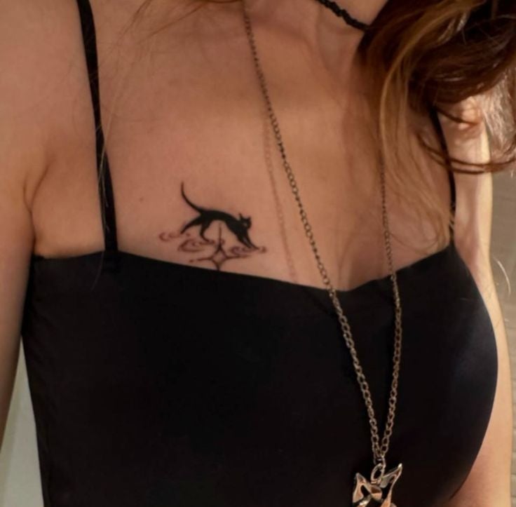Persona con vestido negro de tirantes finos, lleva un collar y tiene un tatuaje de gato negro en el pecho