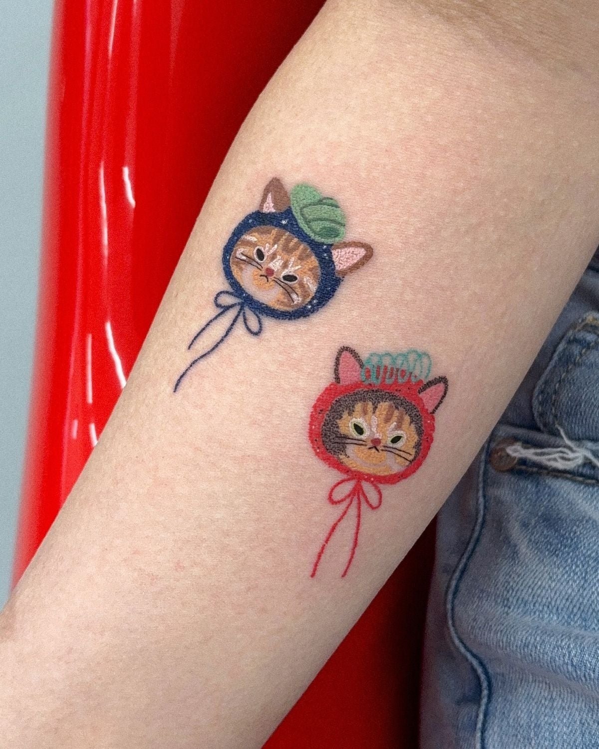 Tatuajes de dos gatos en un brazo, uno con capucha azul y el otro con capucha roja, ambos con lazos