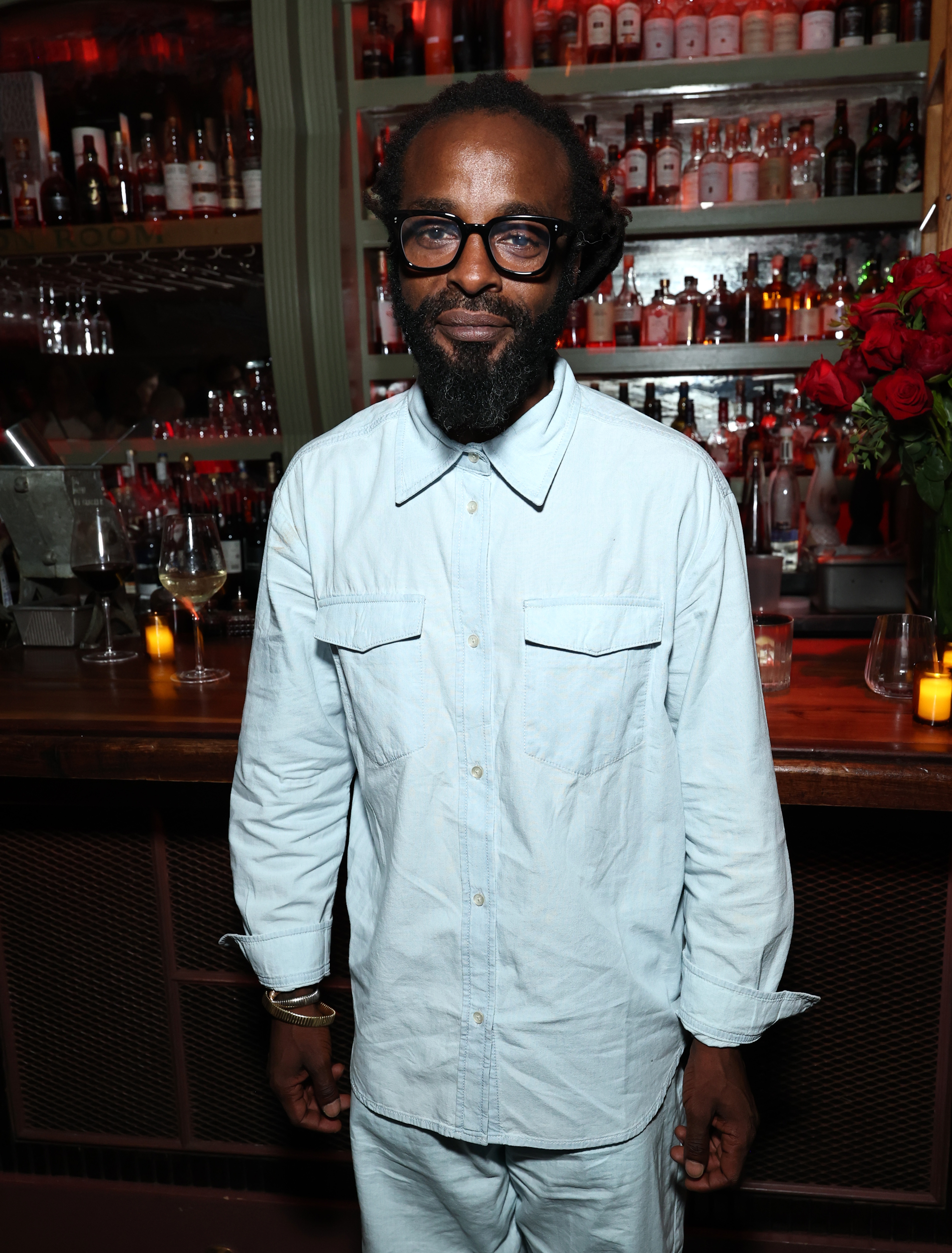 Mann med briller og skjegg iført en lys skjorte med knapper står foran en bar med flasker og blomster