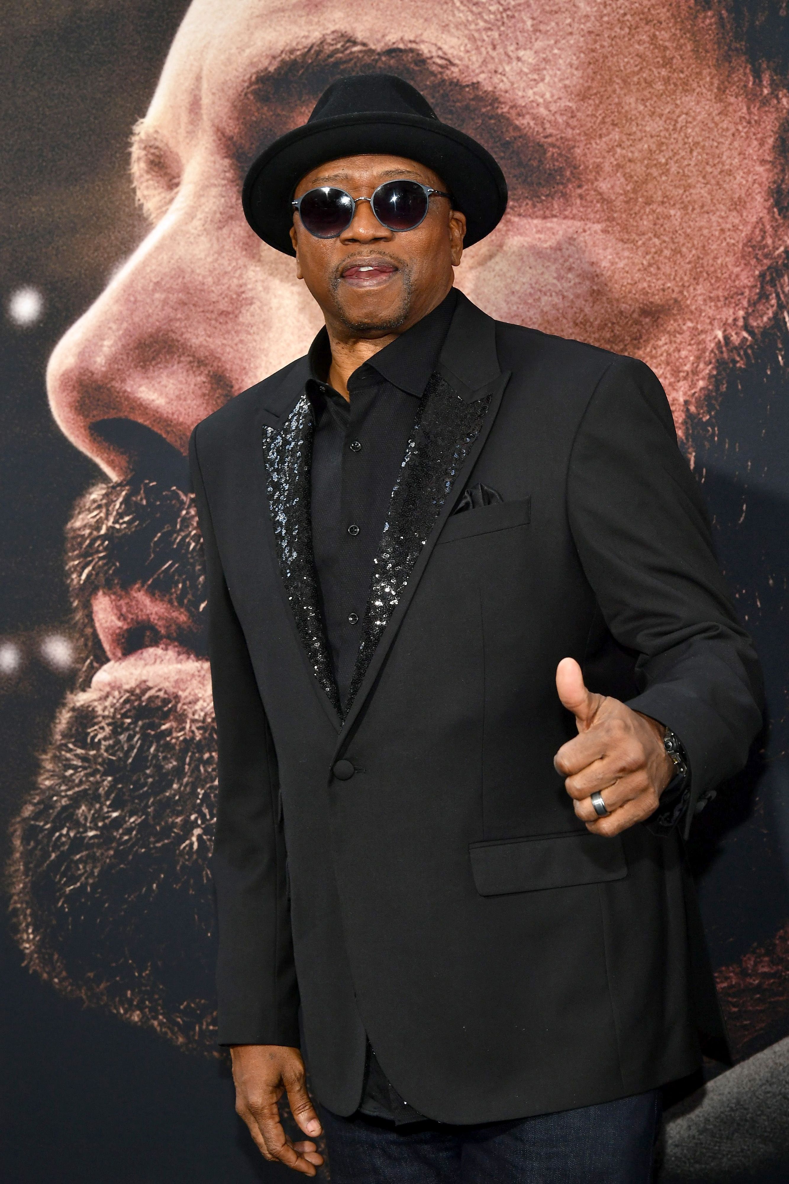 Person i paljettblazer og solbriller på den røde løperen, gir tommel opp, med et stort portrettbilde i bakgrunnen