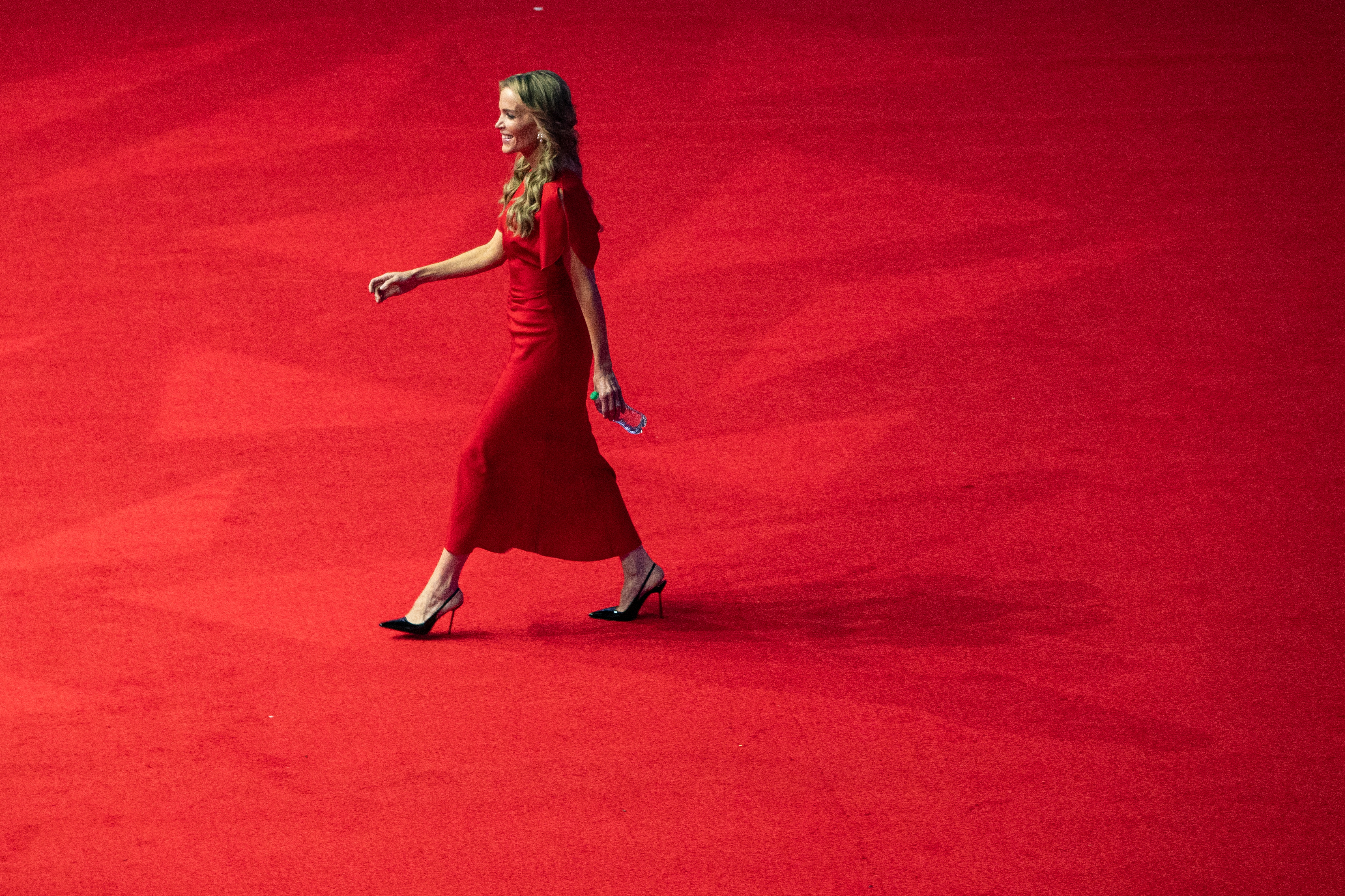 Person successful  elegant, floor-length gown and heels walking connected  a reddish  carpet, holding a clutch