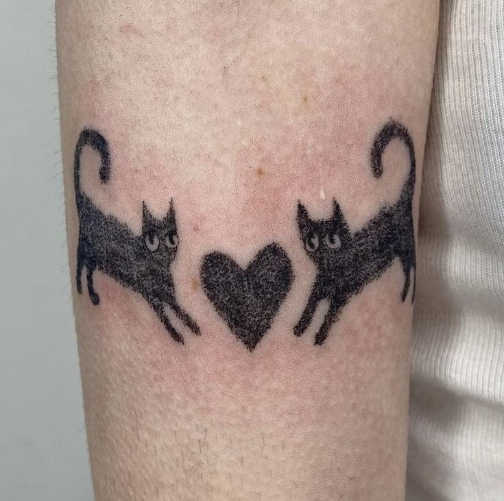 Dos gatos negros estilizados mirándose con un corazón negro entre ellos, tatuados en un brazo