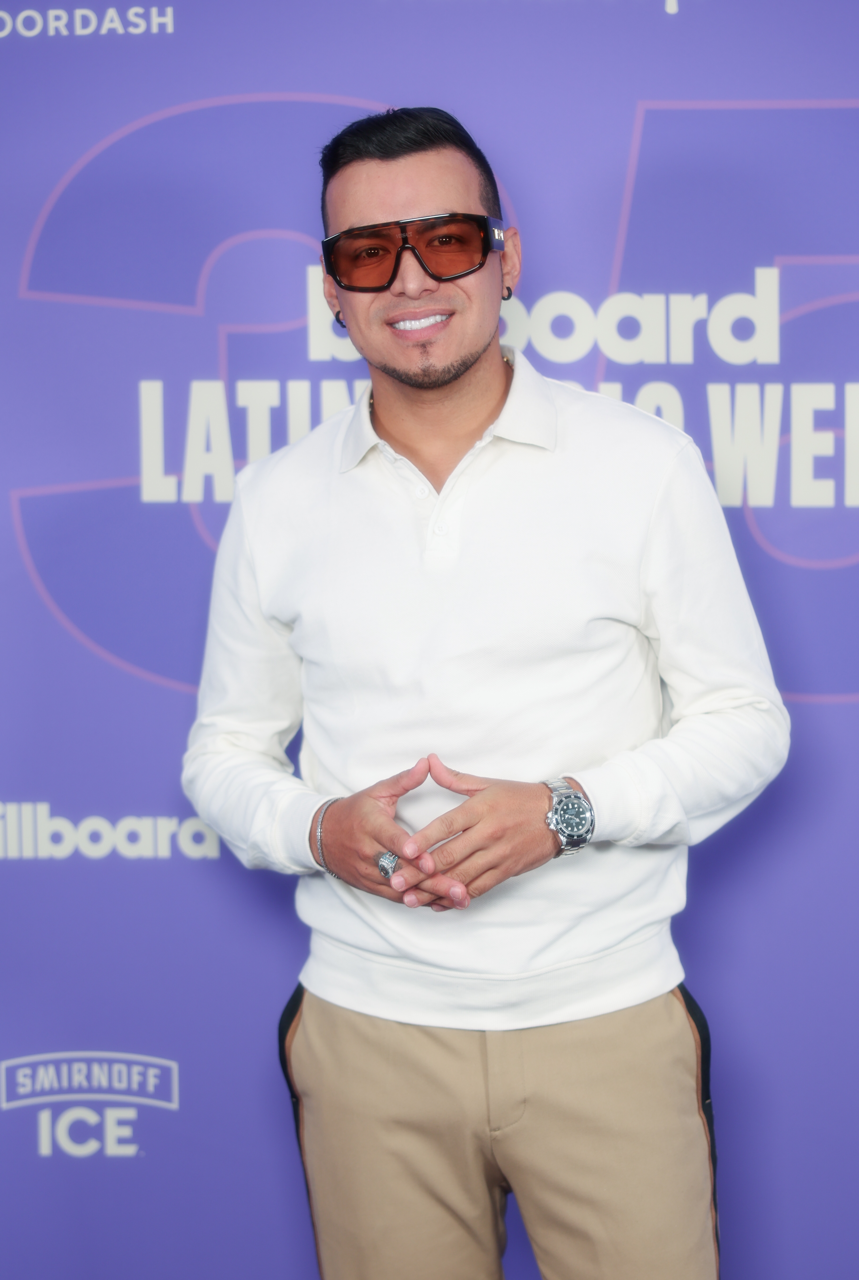 Smilende mann i uformelt antrekk, inkludert brede solbriller og en hvit skjorte, poserer på Billboard Latin Music Week-arrangementet