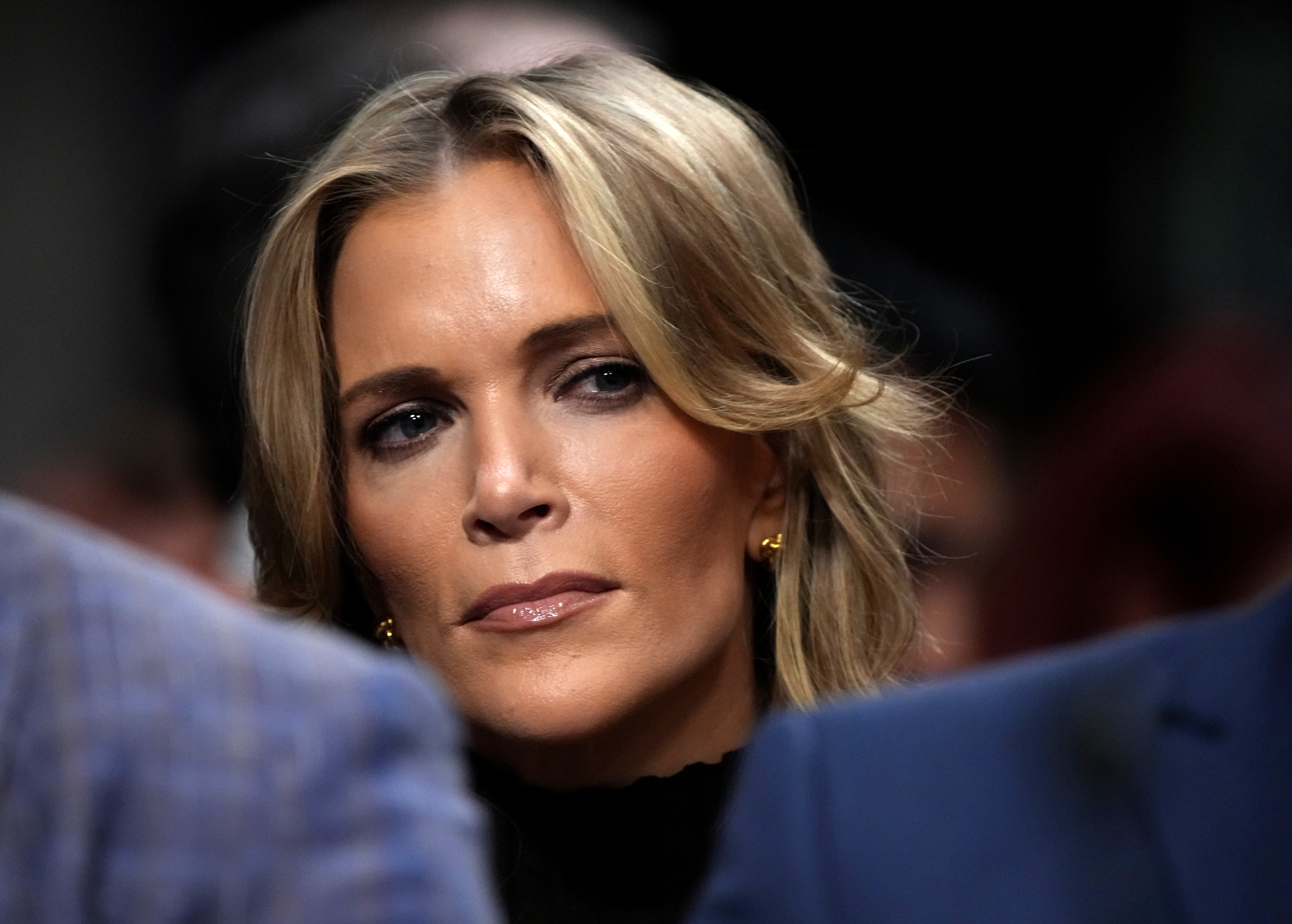 Person with wavy, shoulder-length hairsbreadth  and golden  earrings, wearing a high-neck outfit, looking up  thoughtfully amid a crowd