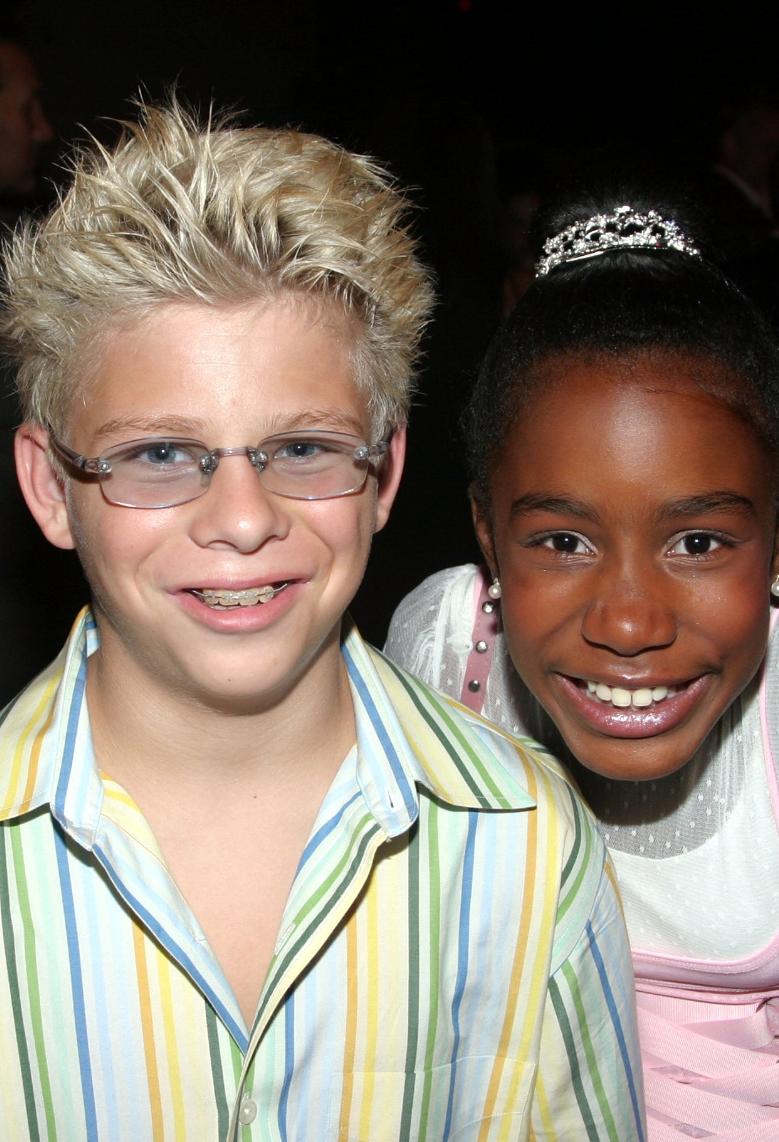 To smilende barn på et arrangement. Gutten har piggete hår og stripet skjorte. Jenta har på seg en diadem og en kjole med et rent overlegg