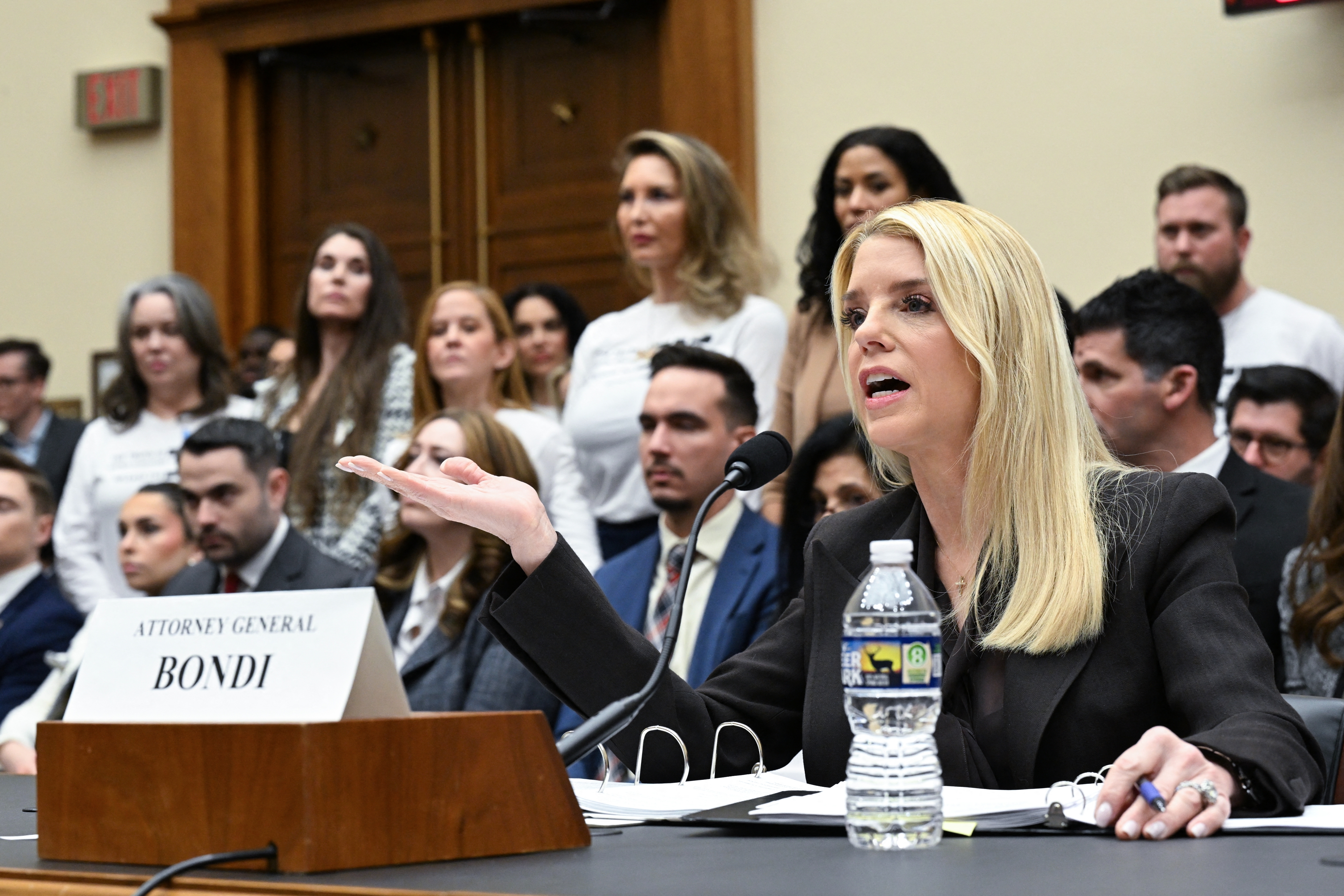 A pistillate   speaks into a microphone astatine  a hearing, surrounded by seated and lasting  people, with a nameplate speechmaking  "Attorney General Bondi."