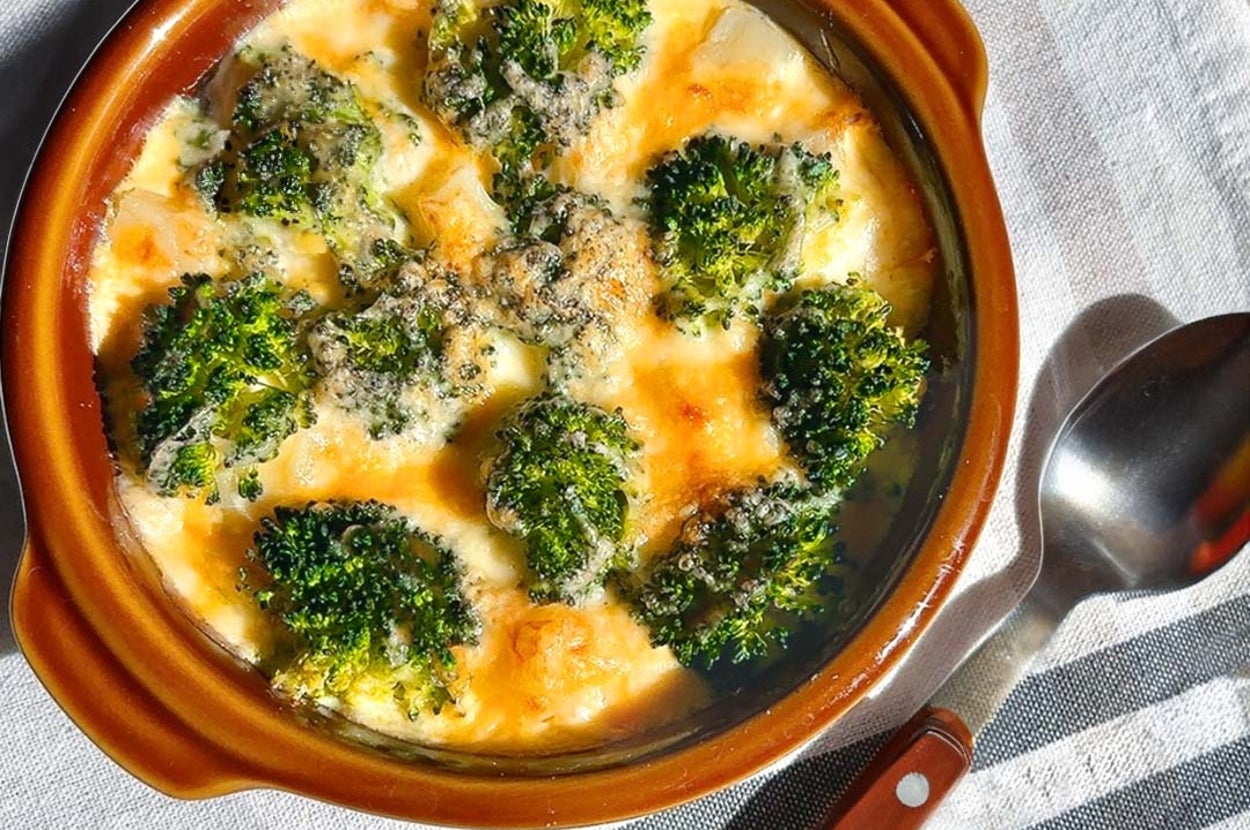 ブロッコリーのグラタンが陶器の皿に盛られ、テーブルクロスの上に置かれている。スプーンが横に添えられている。