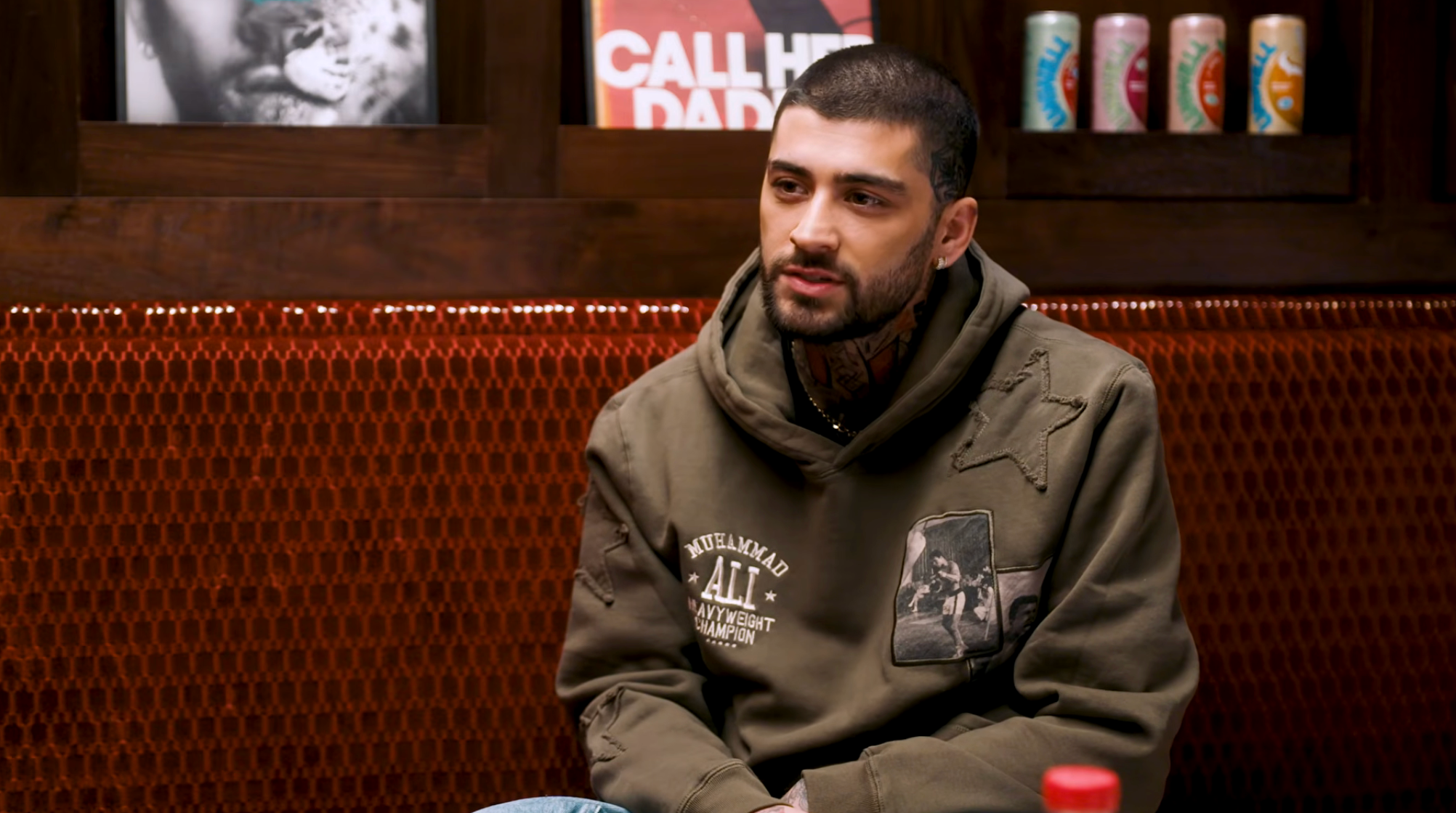 A idiosyncratic   is seated successful  a room, wearing a casual hooded sweatshirt with graphic patches, adjacent  to shelves with posters and beverages