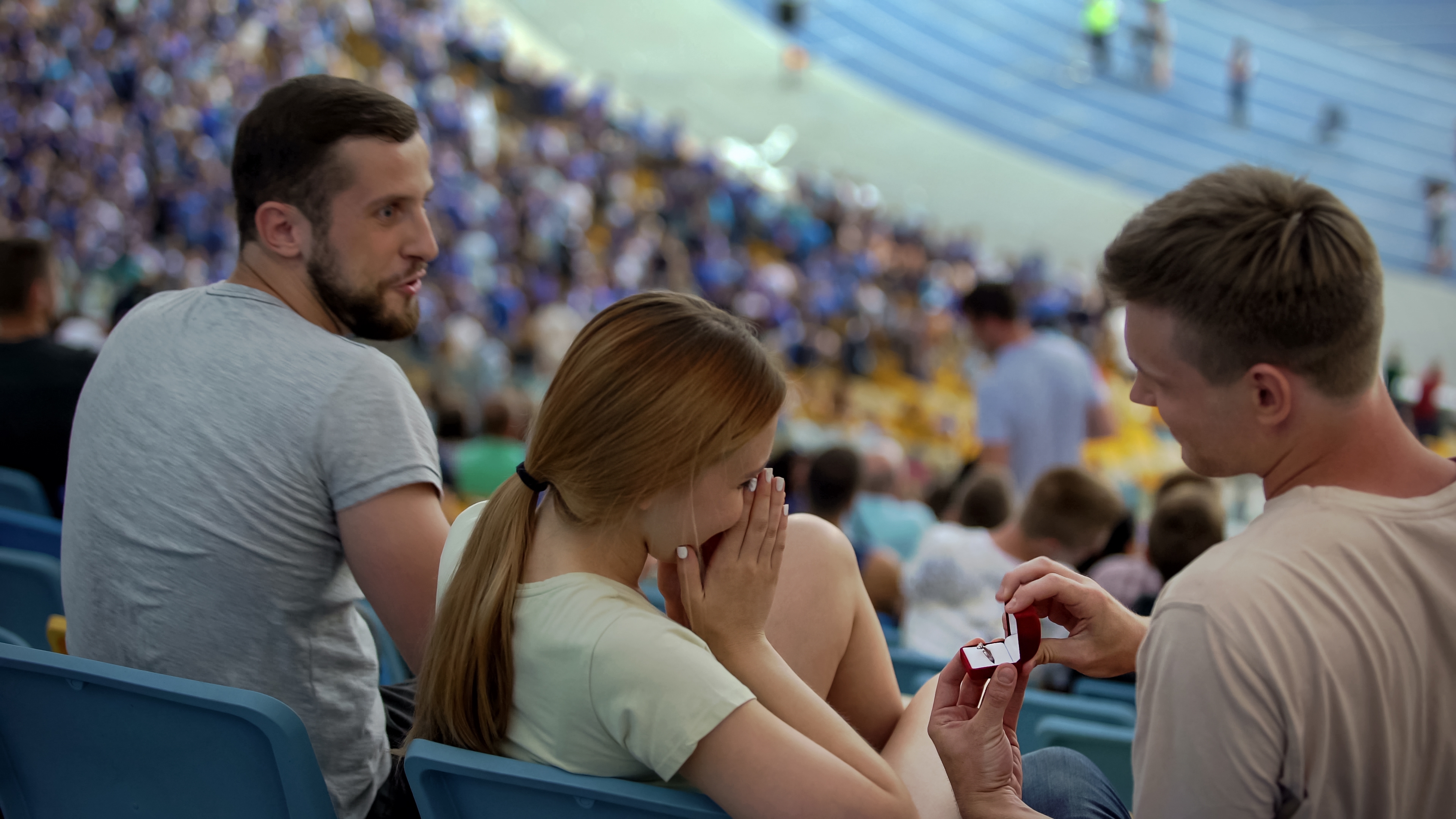 A antheral   successful  stadium seats proposes to a amazed  woman, holding a ring. Another idiosyncratic   watches successful  the background, with a sports lawsuit   occurring