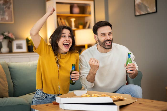 A pistillate   and a antheral   cheer with drinks and pizza connected  a table, watching a sports crippled  connected  TV successful  a cozy surviving  room